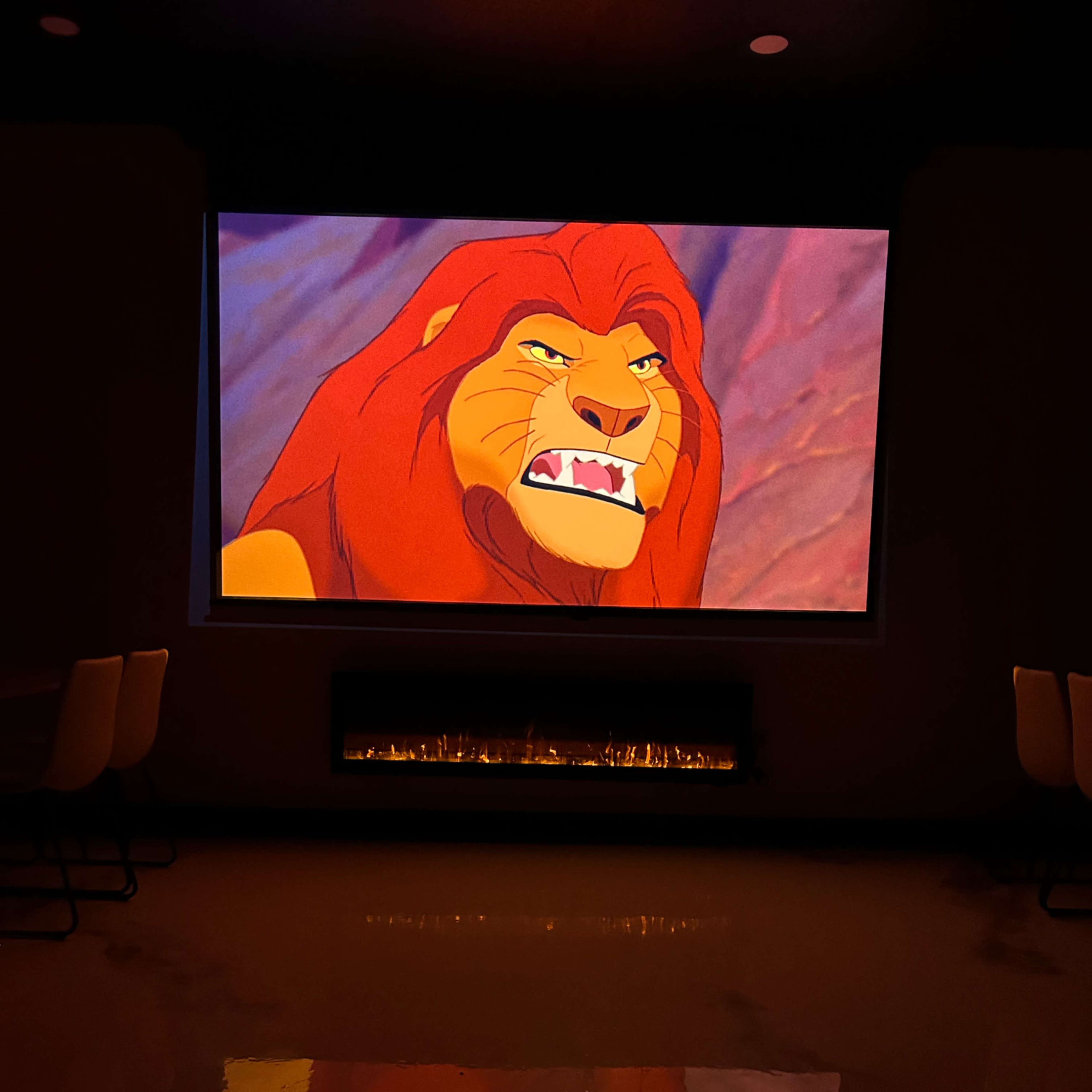 A large screen displays an animated lion character, while a fireplace is visible below it in a dimly lit room.