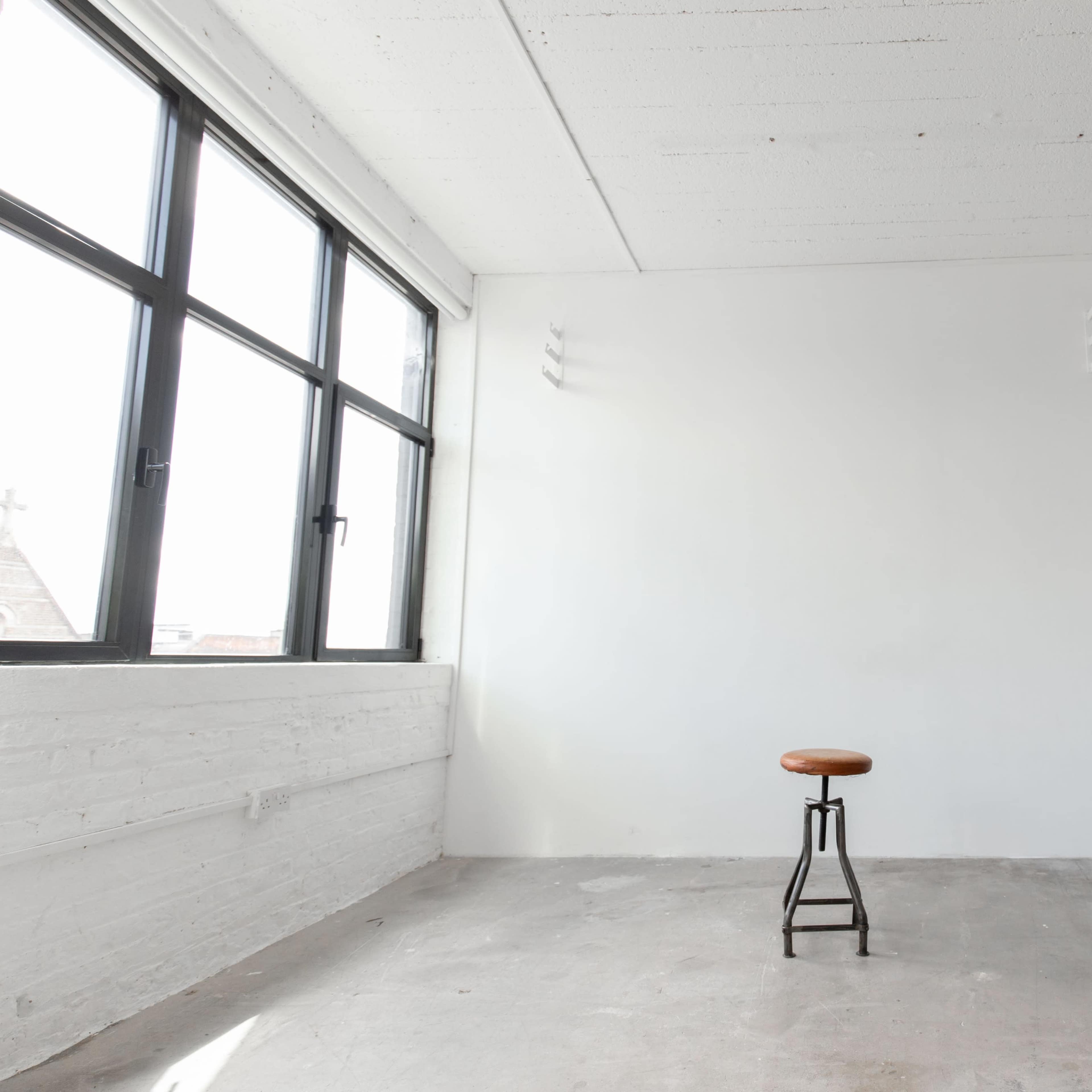 A minimalist interior space with a single wooden stool positioned in front of a blank white wall, large windows, and a potted plant in the corner.