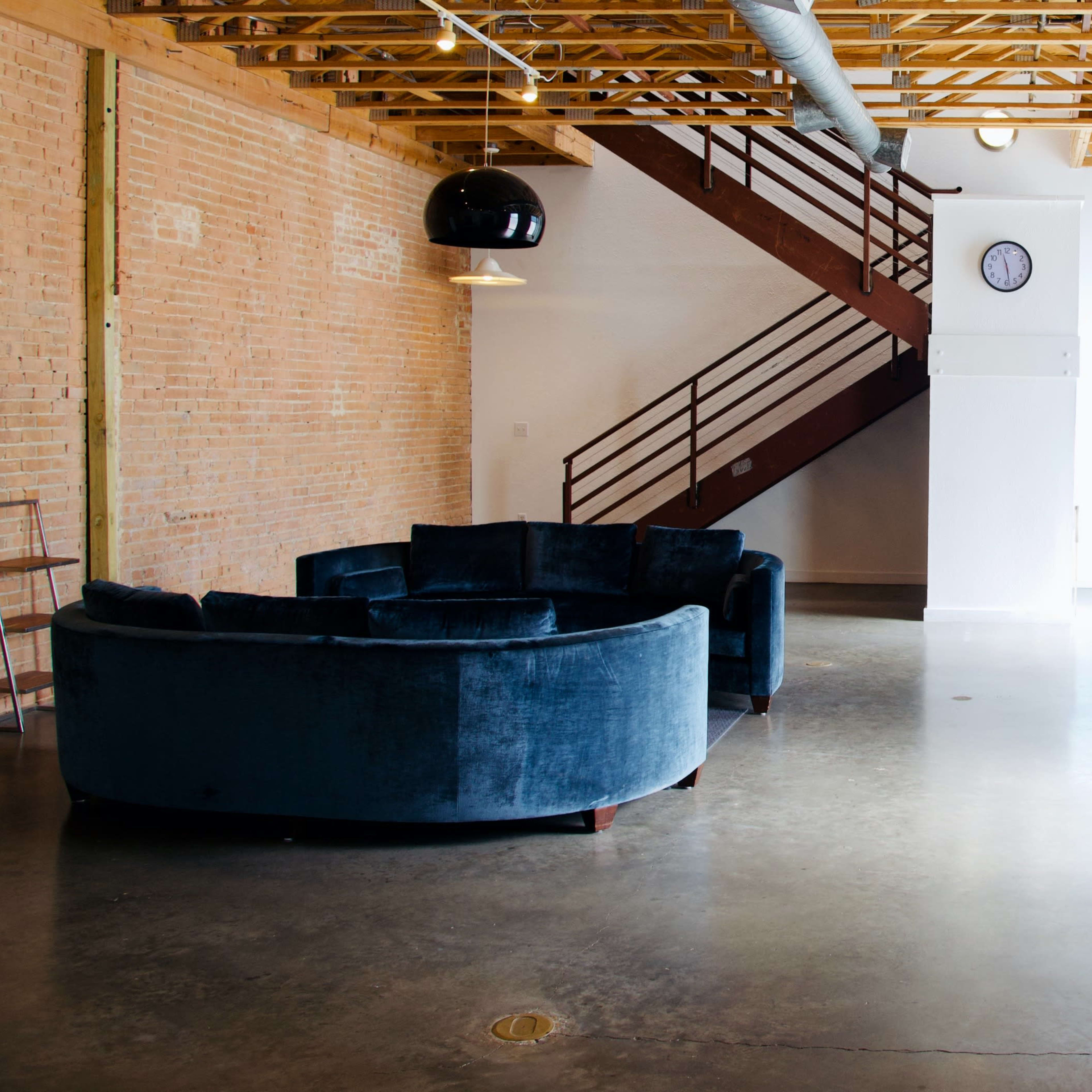 A spacious interior with exposed brick walls, a circular blue velvet couch, a wooden staircase, and large windows allowing light to fill the area.