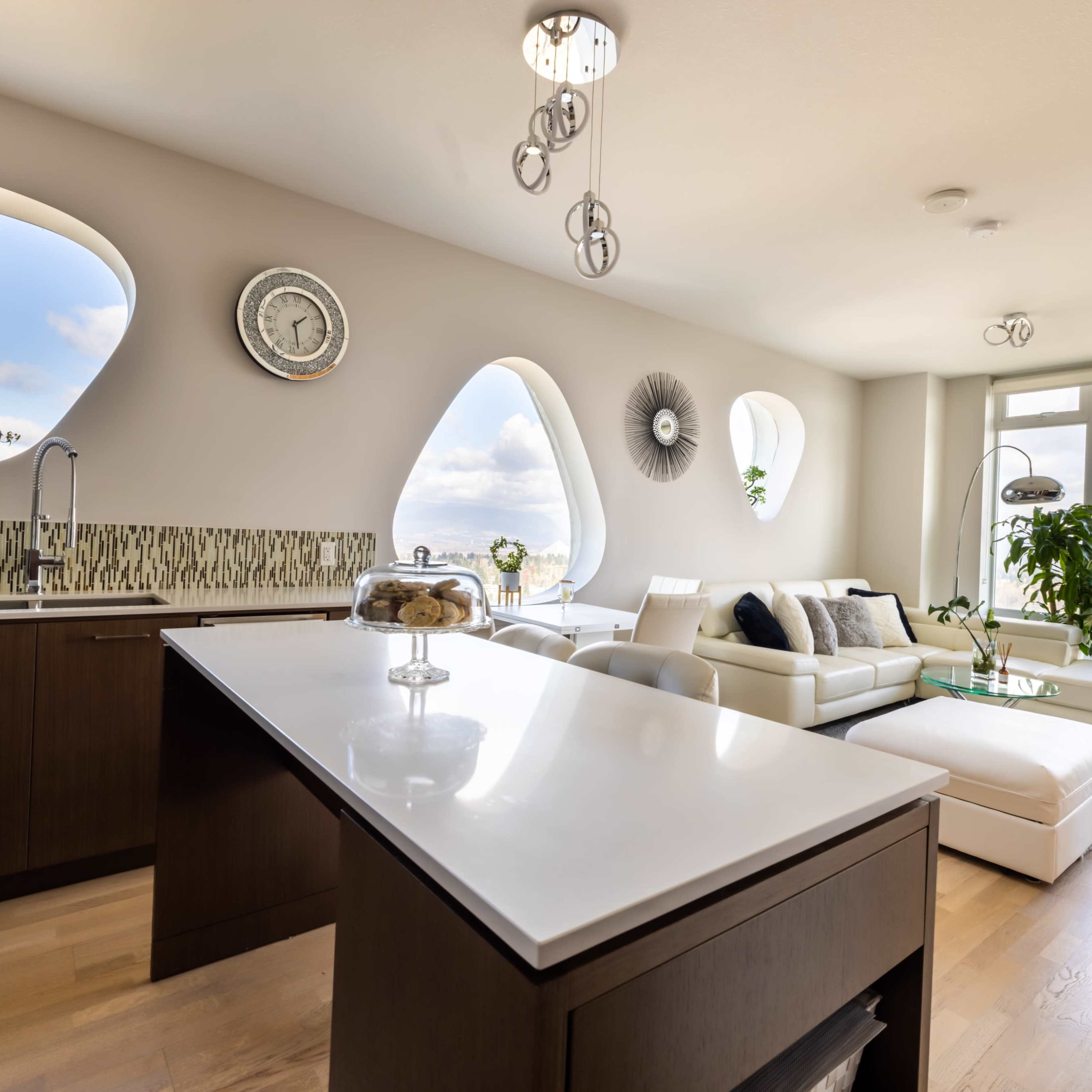 A modern kitchen and living area feature large, uniquely shaped windows, sleek cabinetry, and contemporary furnishings.