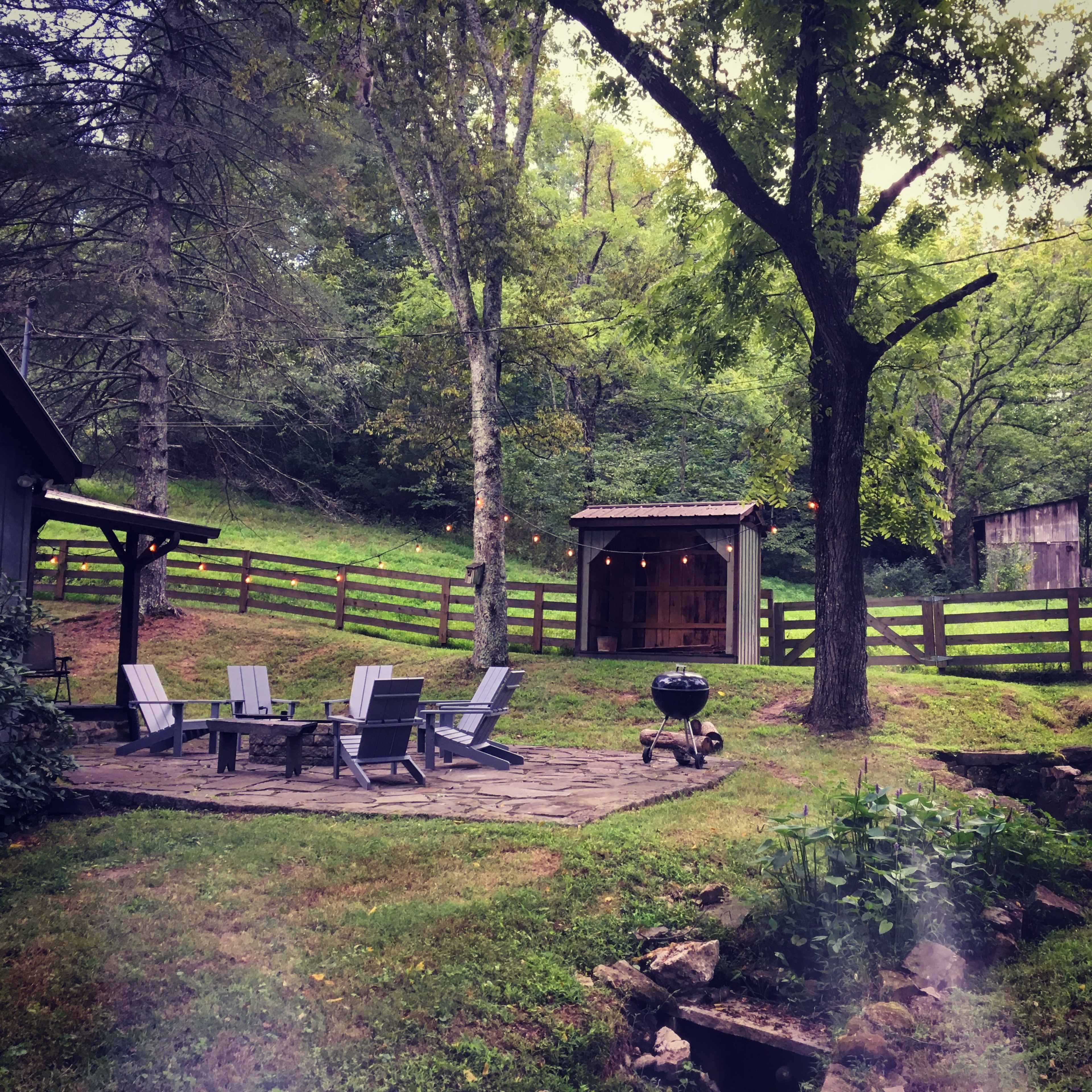 A backyard area with a stone patio surrounded by wooden chairs and a grill, set against a backdrop of trees and a fenced garden.