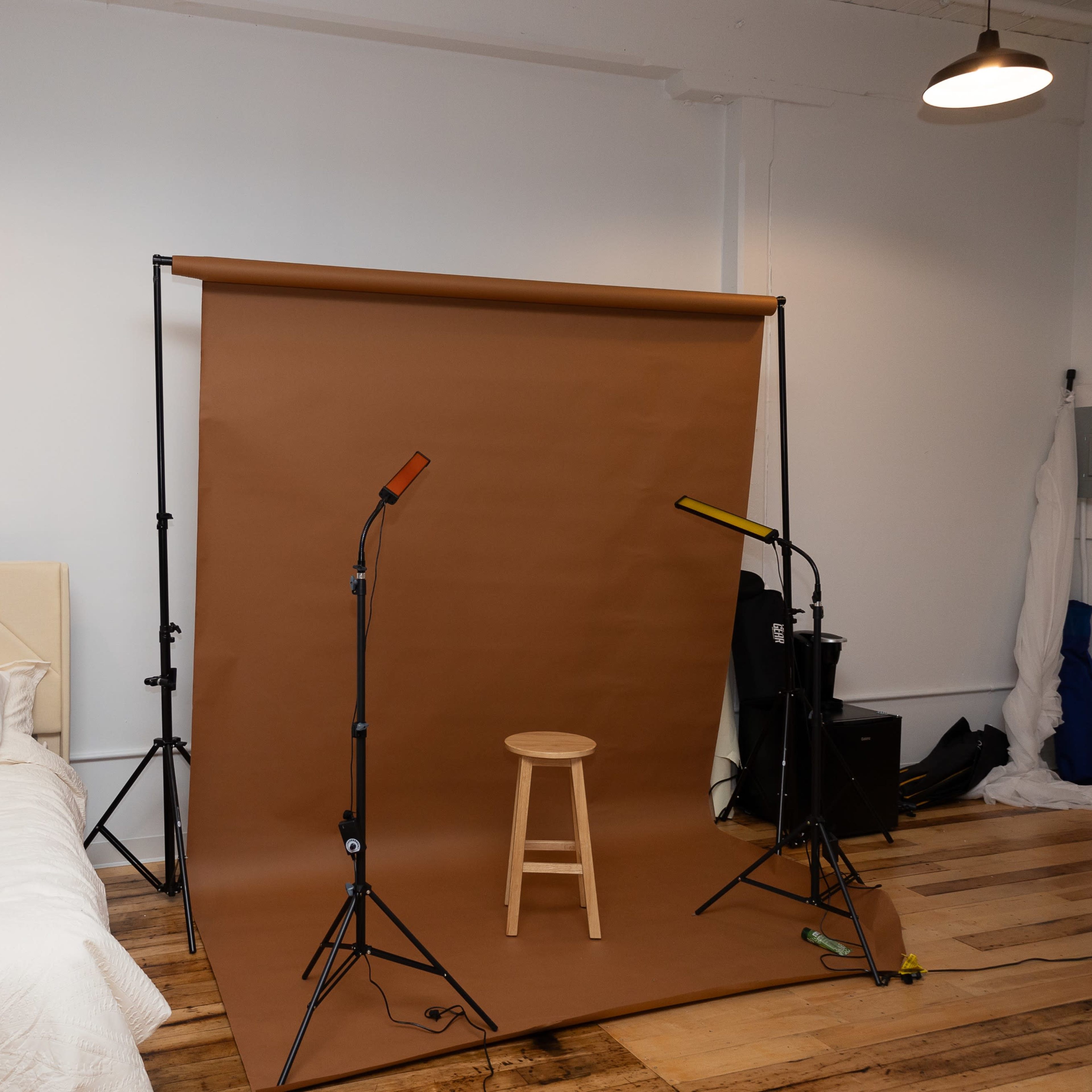 A simple studio setup featuring a bed, a brown backdrop on a stand, and two lighting fixtures positioned around a wooden stool.