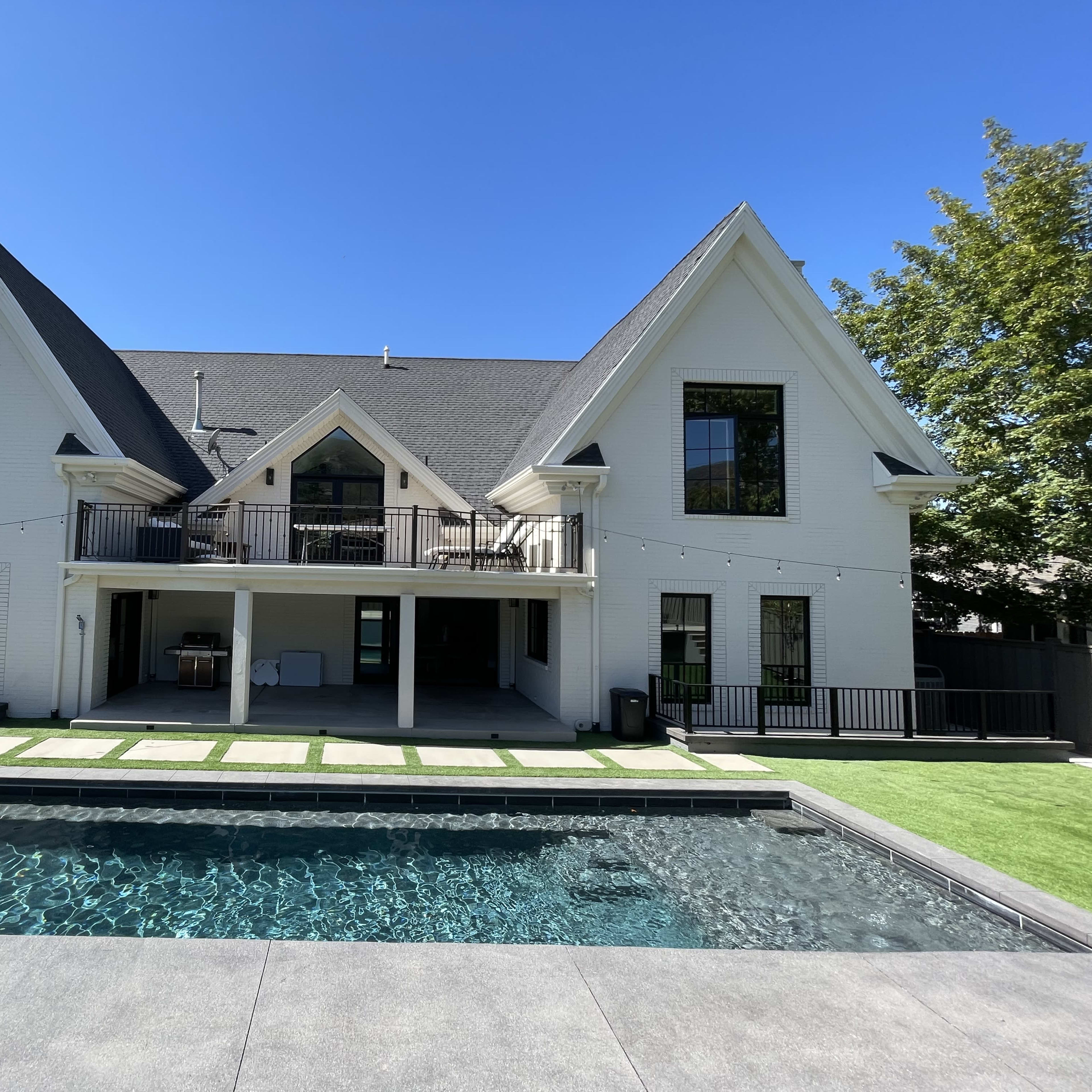 A modern two-story house with a sloped roof and large windows overlooks a rectangular pool surrounded by a stone patio and grass lawn.