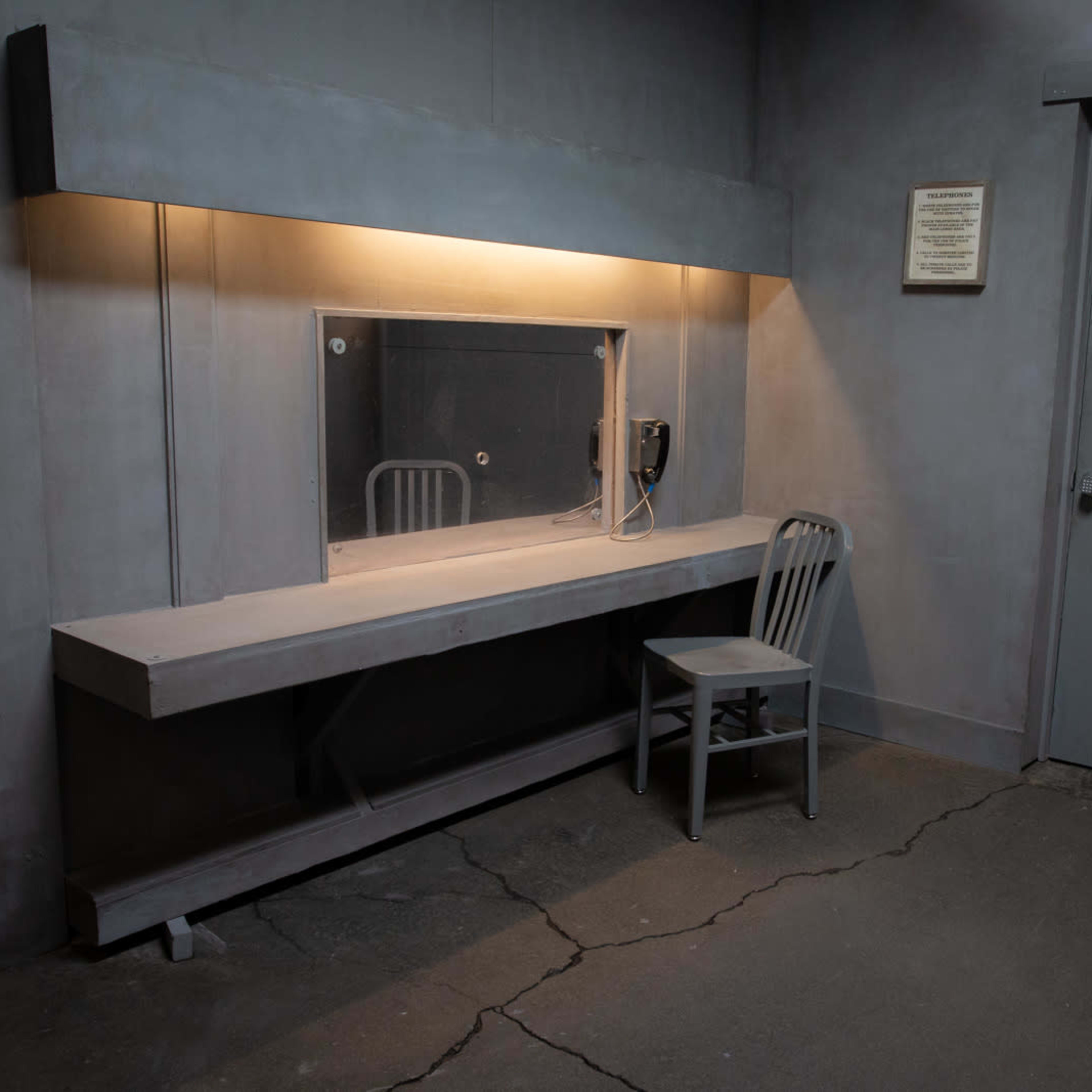 A sparse, gray room with a single chair and a wall-mounted mirror above a long, flat surface, next to a door and a phone.