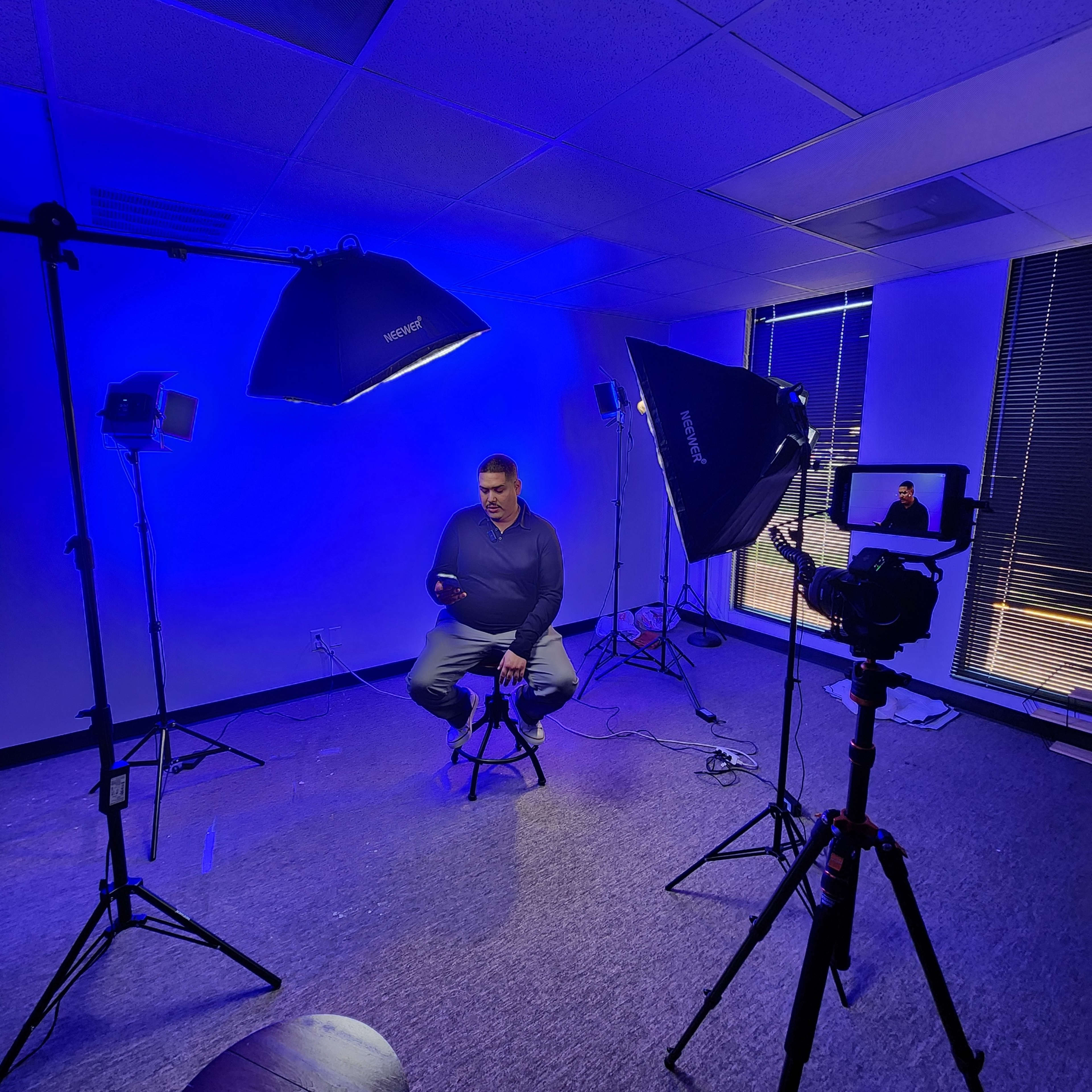 A man sits on a stool in a studio setup with various lighting equipment and a camera focused on him.