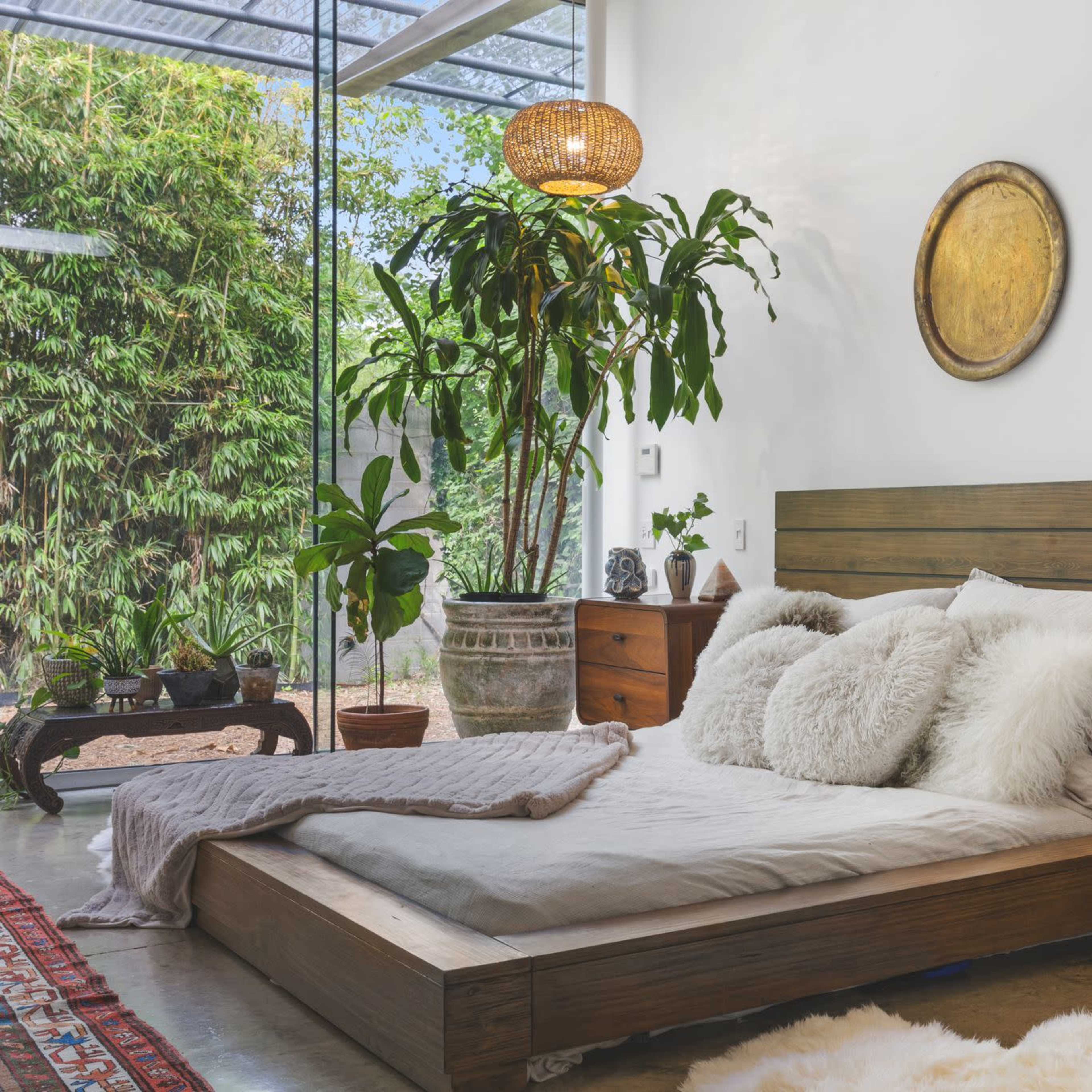 A modern bedroom features a low wooden bed with plush pillows, surrounded by plants and wooden furniture, illuminated by a pendant lamp.