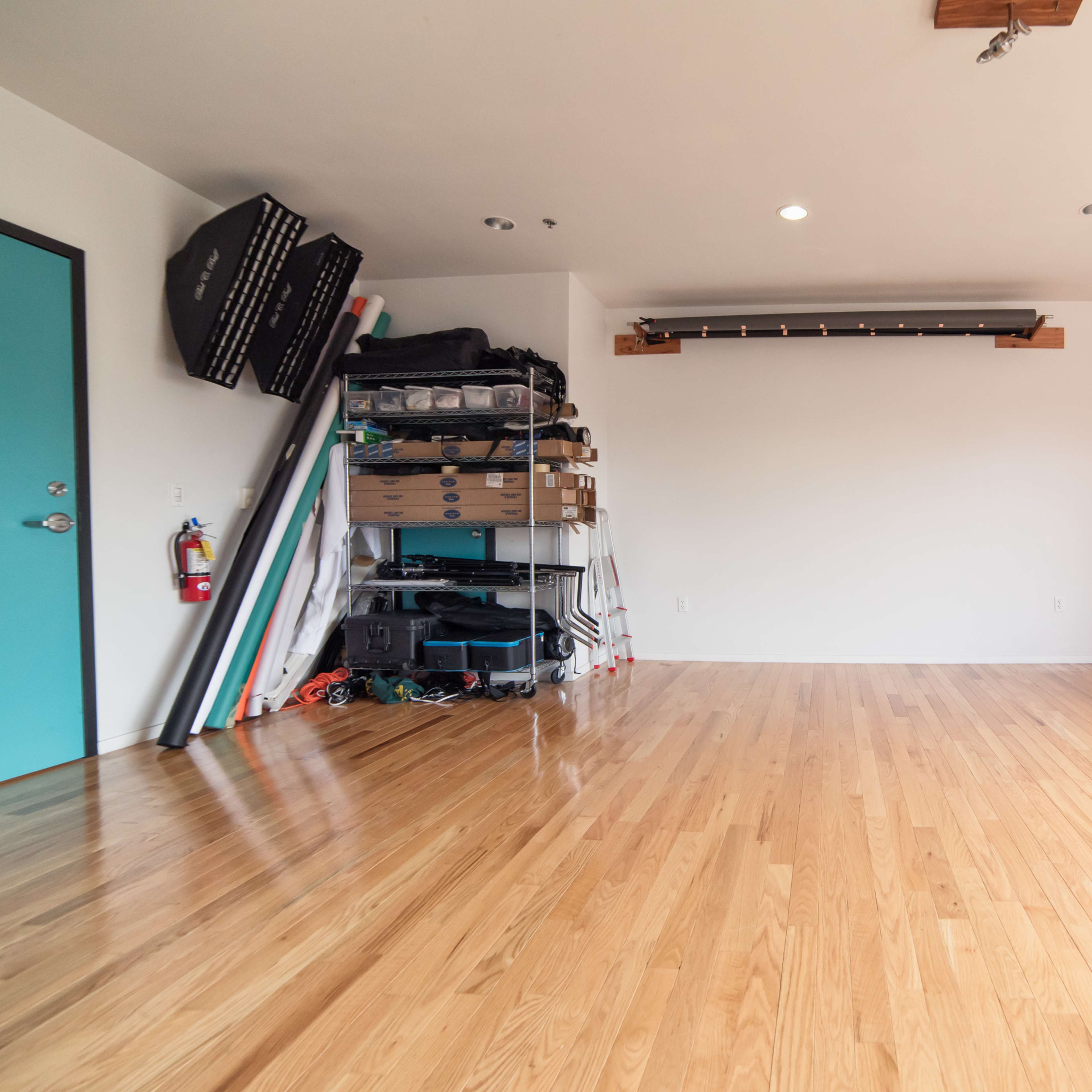 A spacious, empty room featuring light wooden flooring, turquoise doors, and storage shelves with various items along one wall.