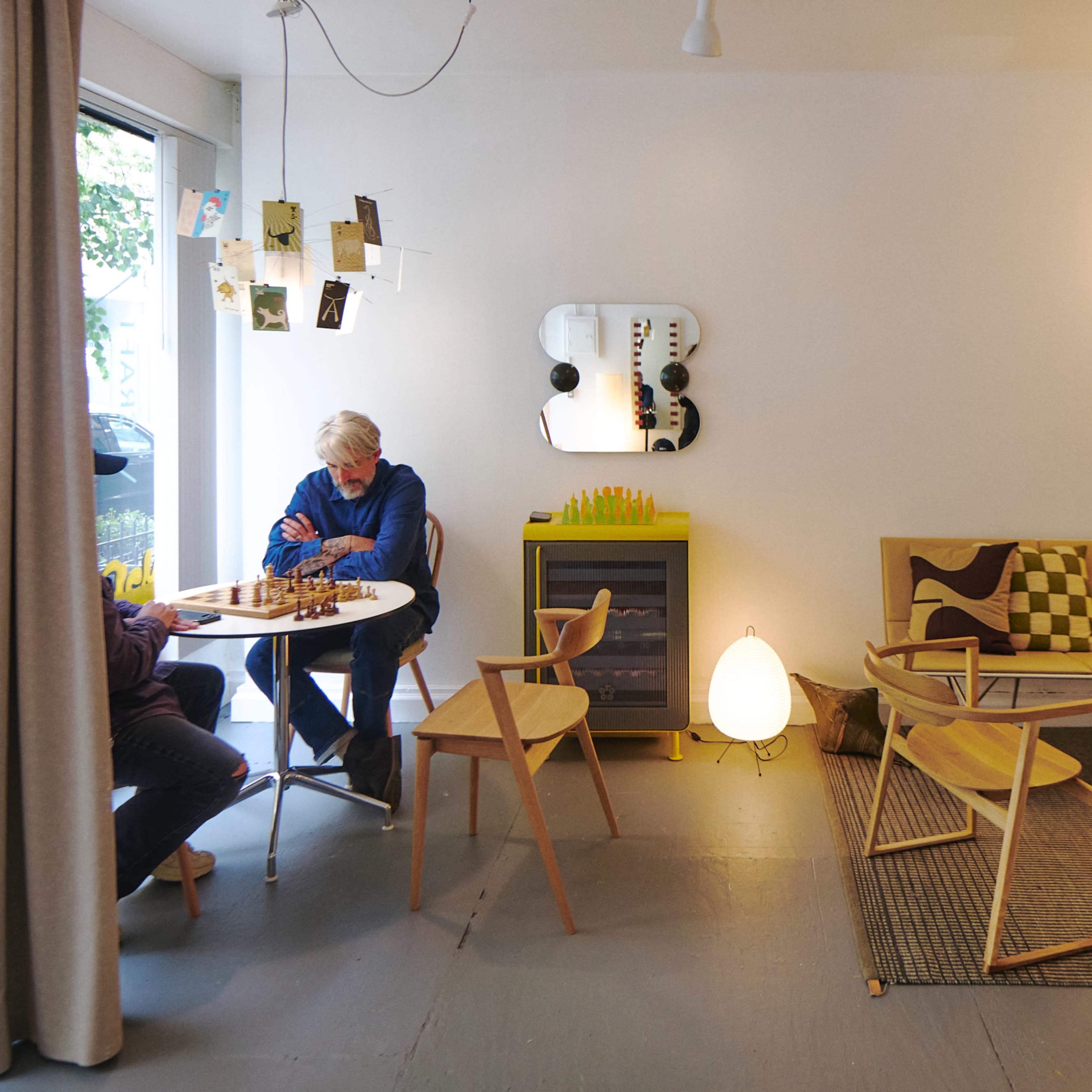 A modern, well-lit room features two people playing chess at a round table, surrounded by minimalist furniture and decorative elements.