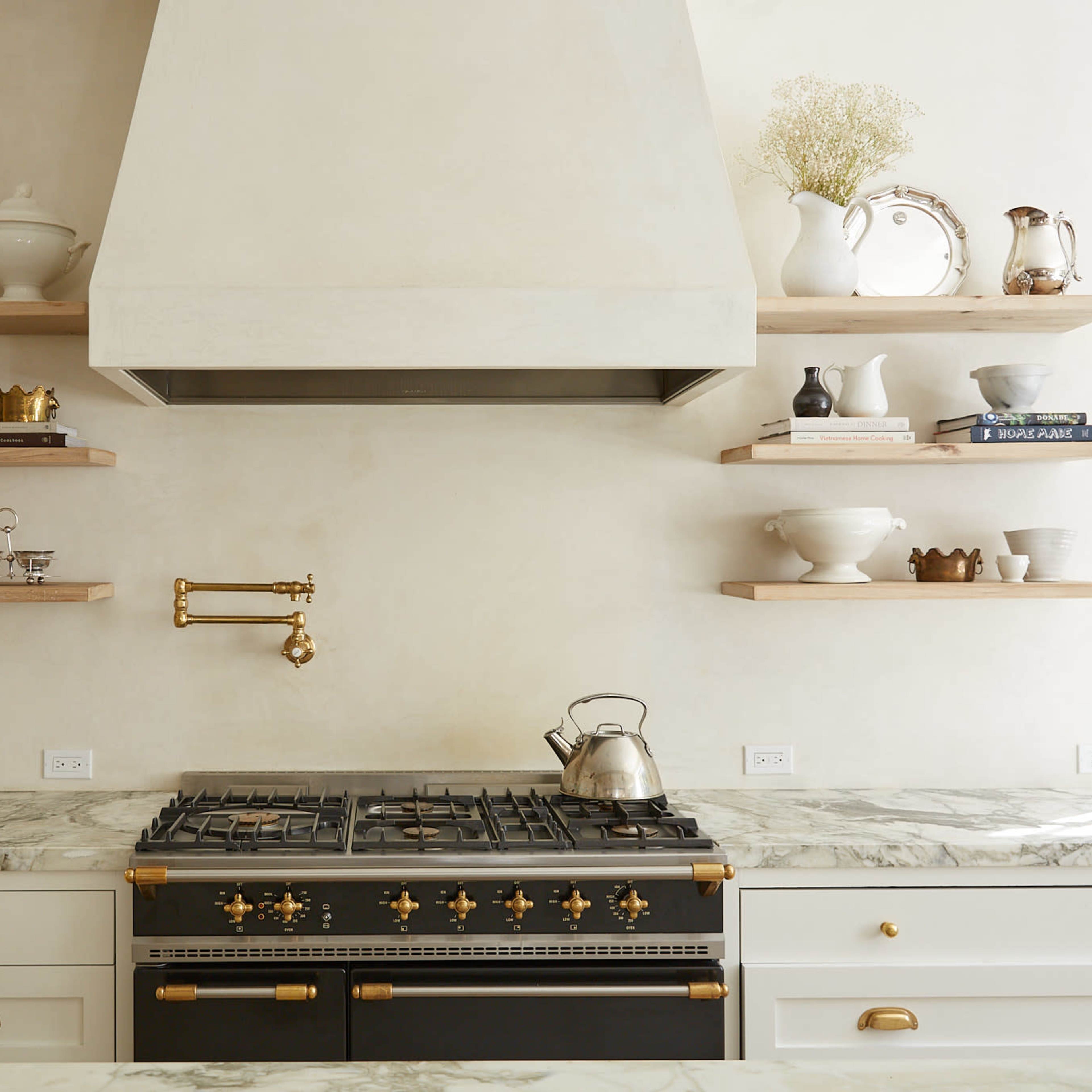 A modern kitchen with a marble countertop, a black and gold stove, and open wooden shelves displaying white dishware.