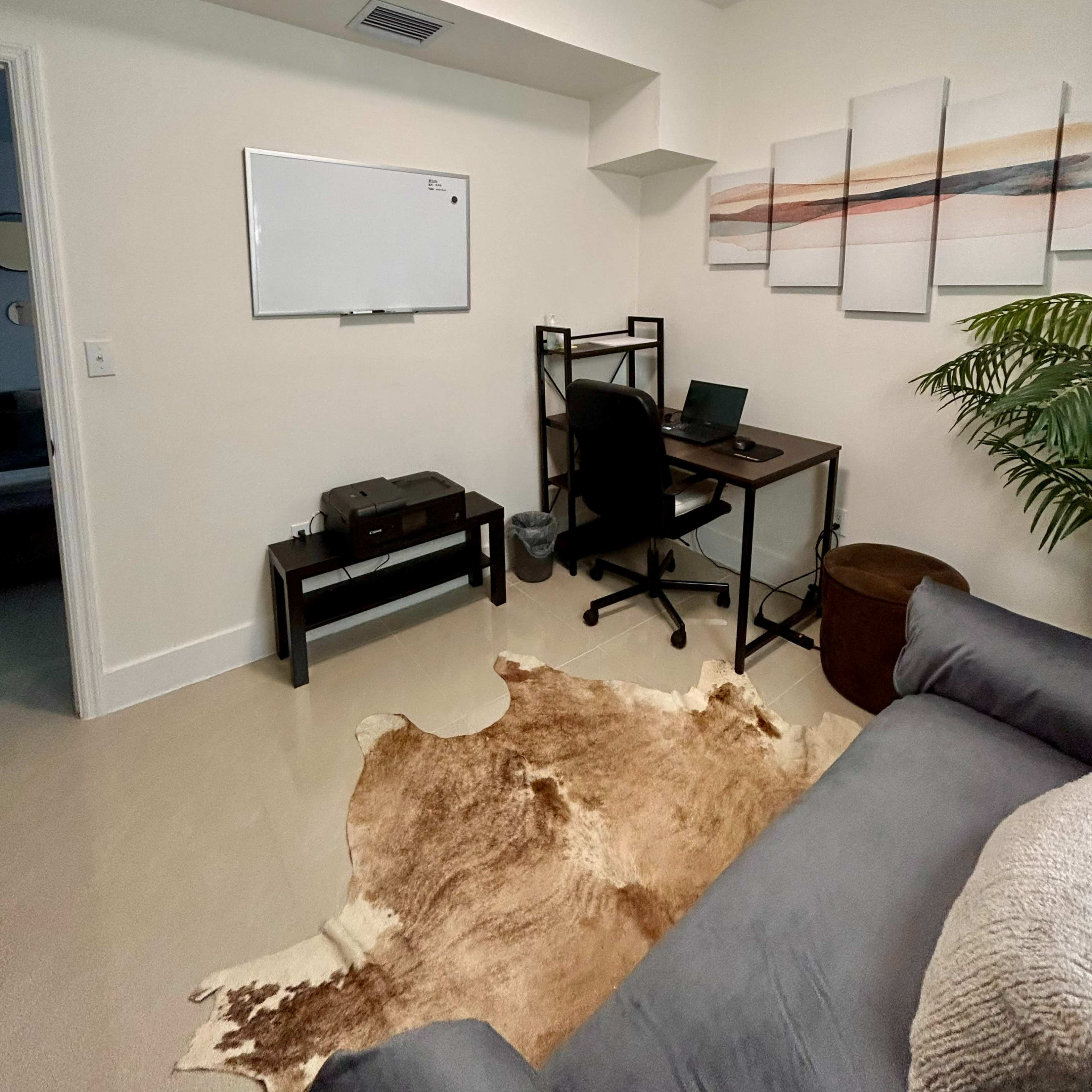 A workspace in a small room featuring a desk with a laptop, a whiteboard on the wall, and a cowhide rug on the floor.