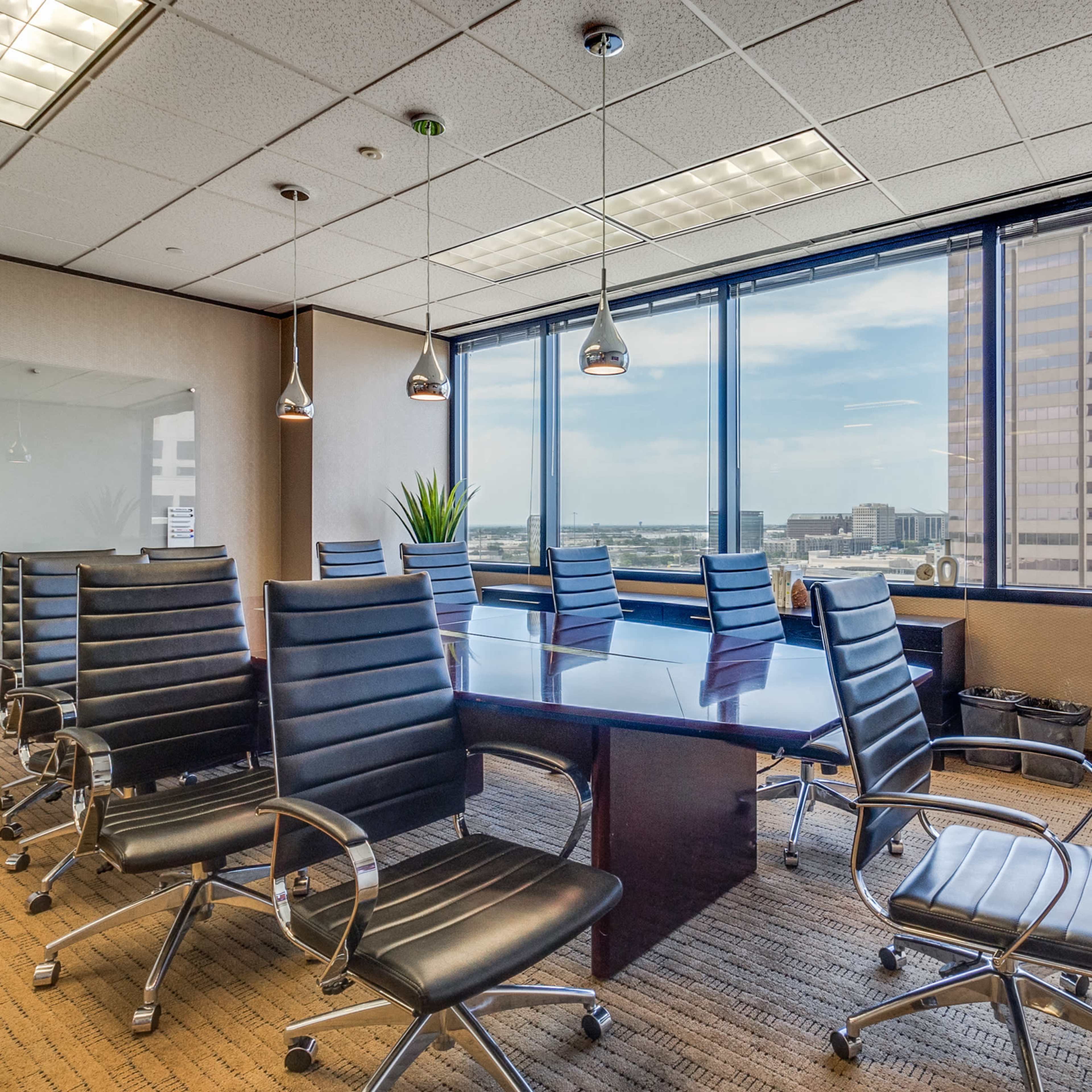 A modern conference room features a large wooden table surrounded by black leather chairs, with windows providing a view of the city skyline.