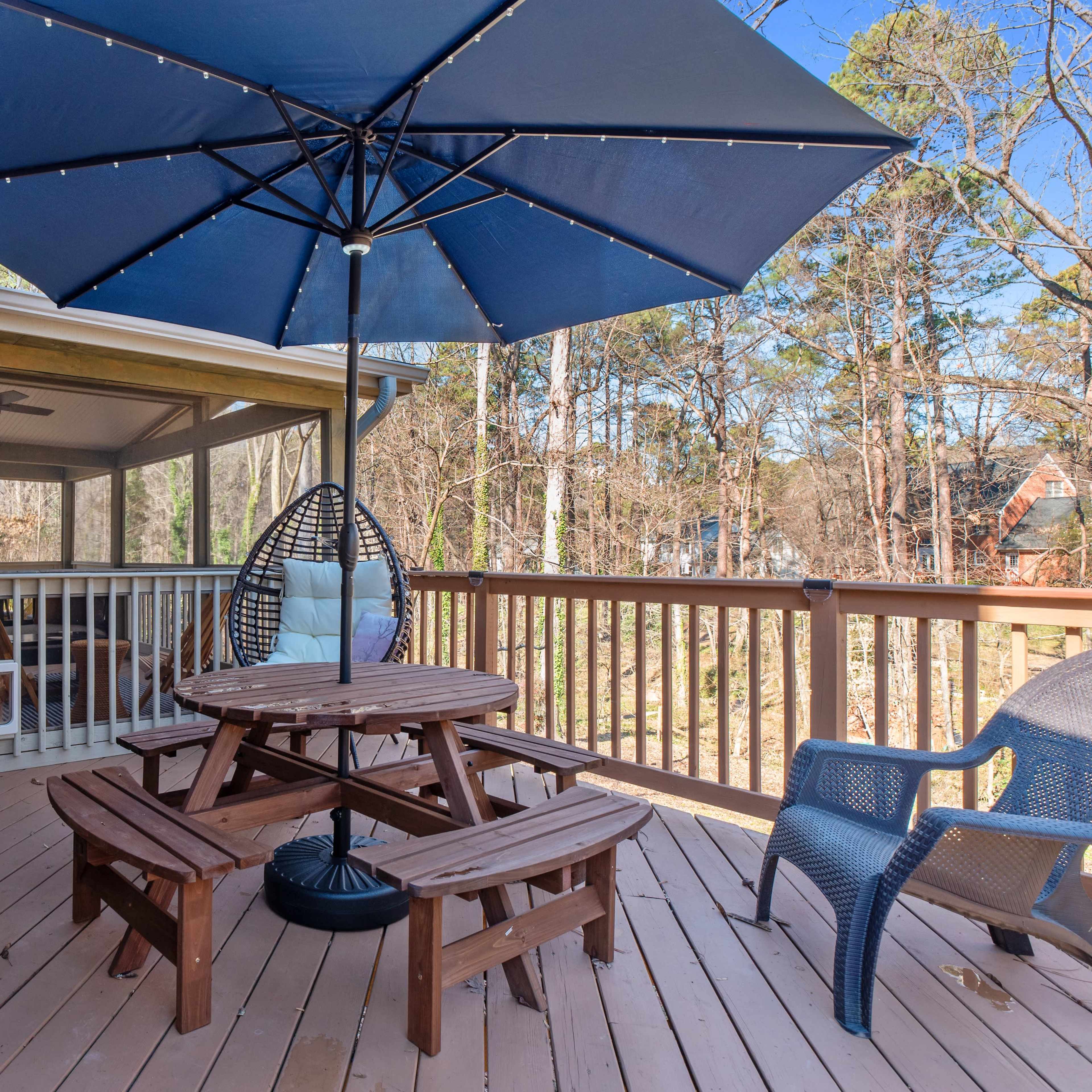 A wooden deck features a round picnic table with benches, an umbrella, and a single chair, surrounded by trees.