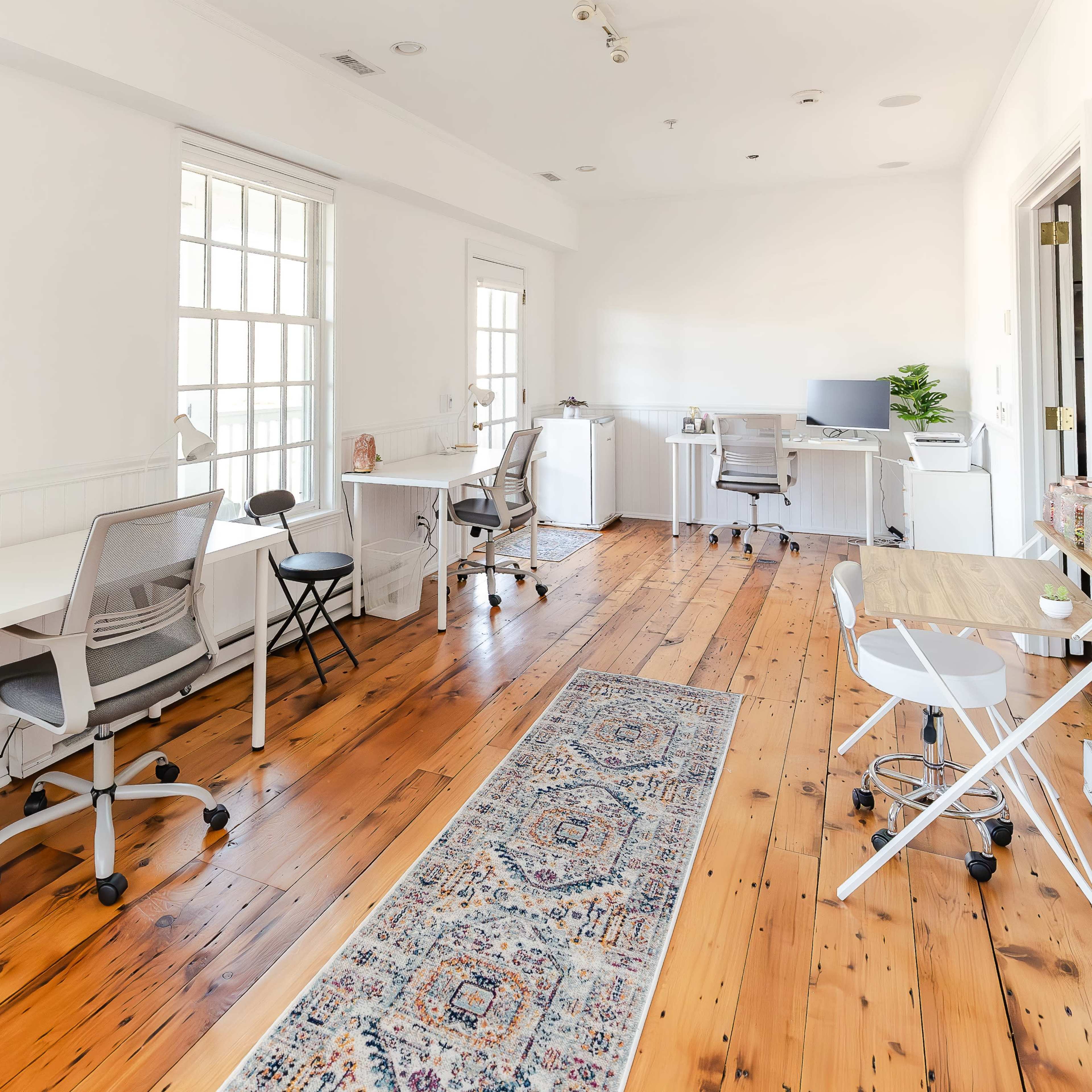 A bright, spacious office with wooden flooring, three workstations, and large windows allowing natural light to fill the room.