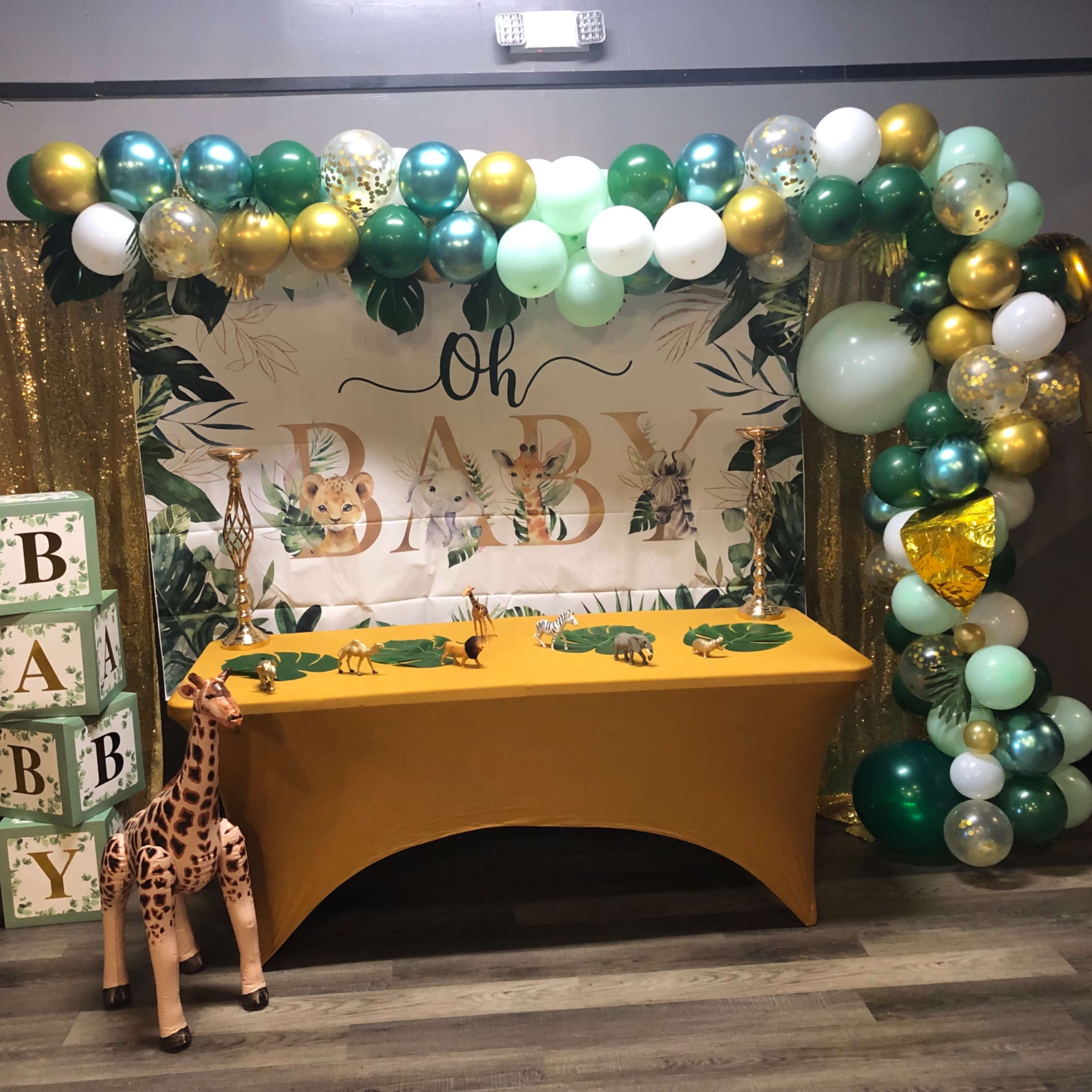 A decorative baby shower setup featuring a table with a yellow tablecloth, a banner that says "Oh Baby," and colorful balloons in green, gold, and white, along with toy animals placed on the table.