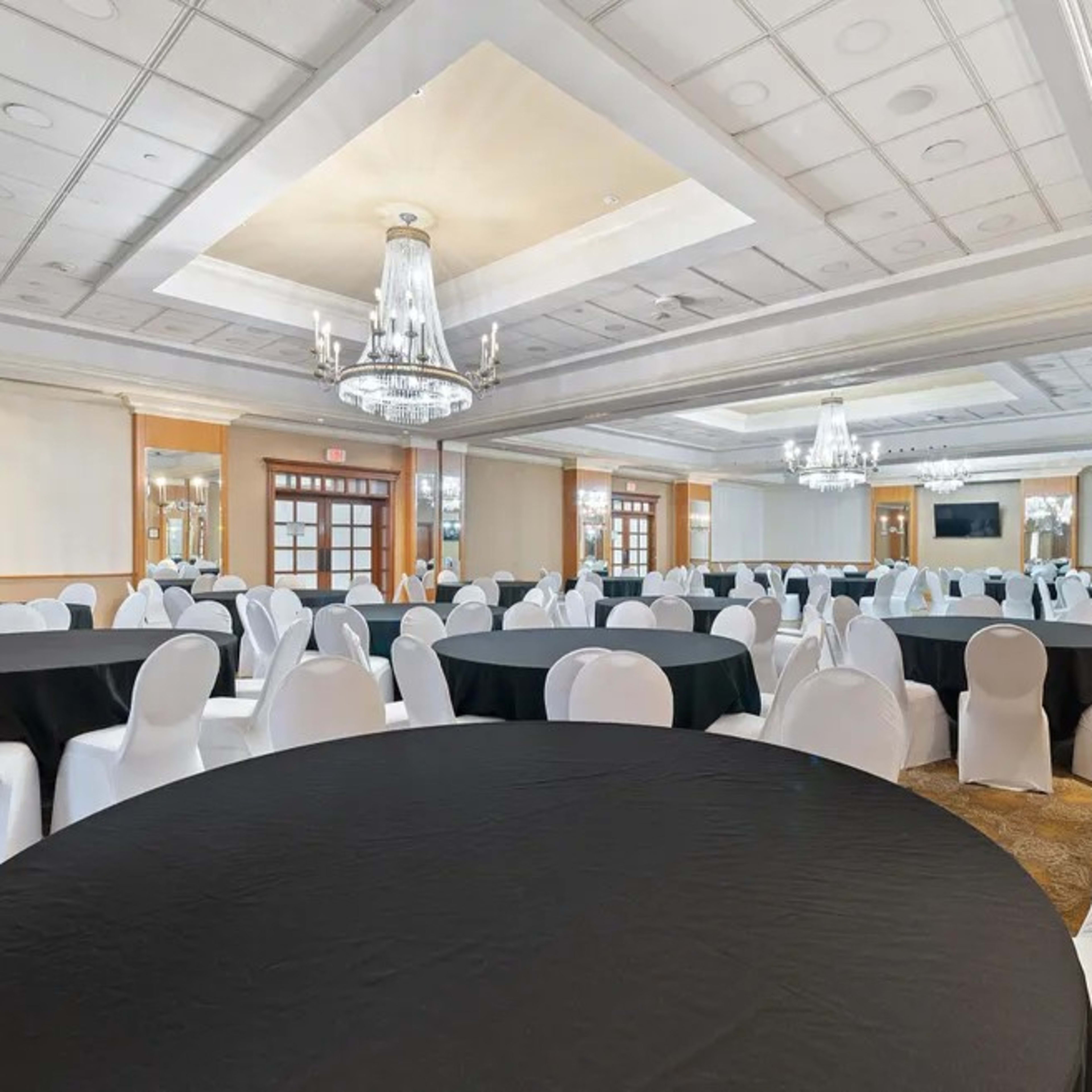 A large event space features multiple round tables covered with black and white tablecloths, all arranged in rows with chandeliers overhead.