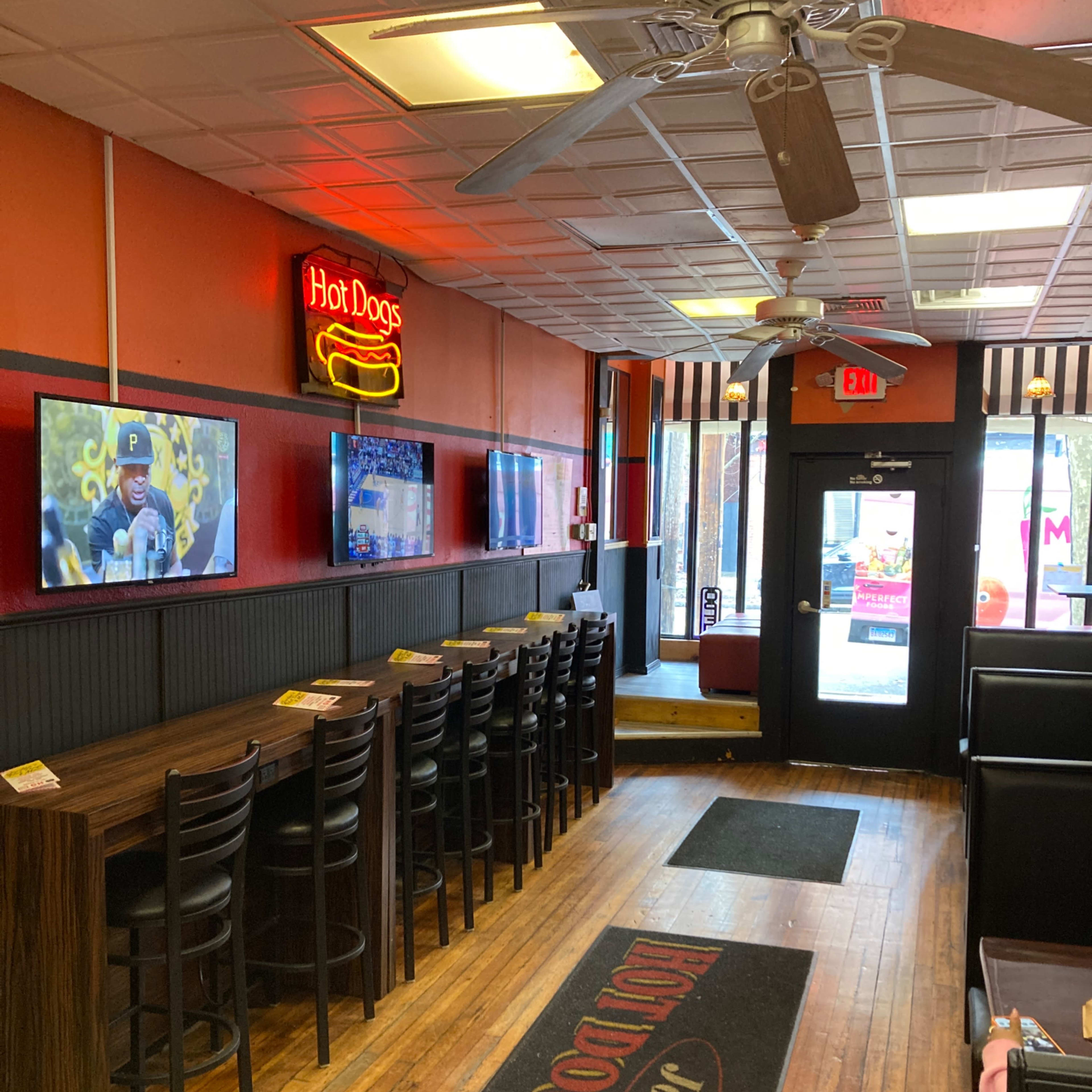 The interior of a hot dog restaurant with a row of bar seating, multiple TVs on the walls, and a door leading outside.