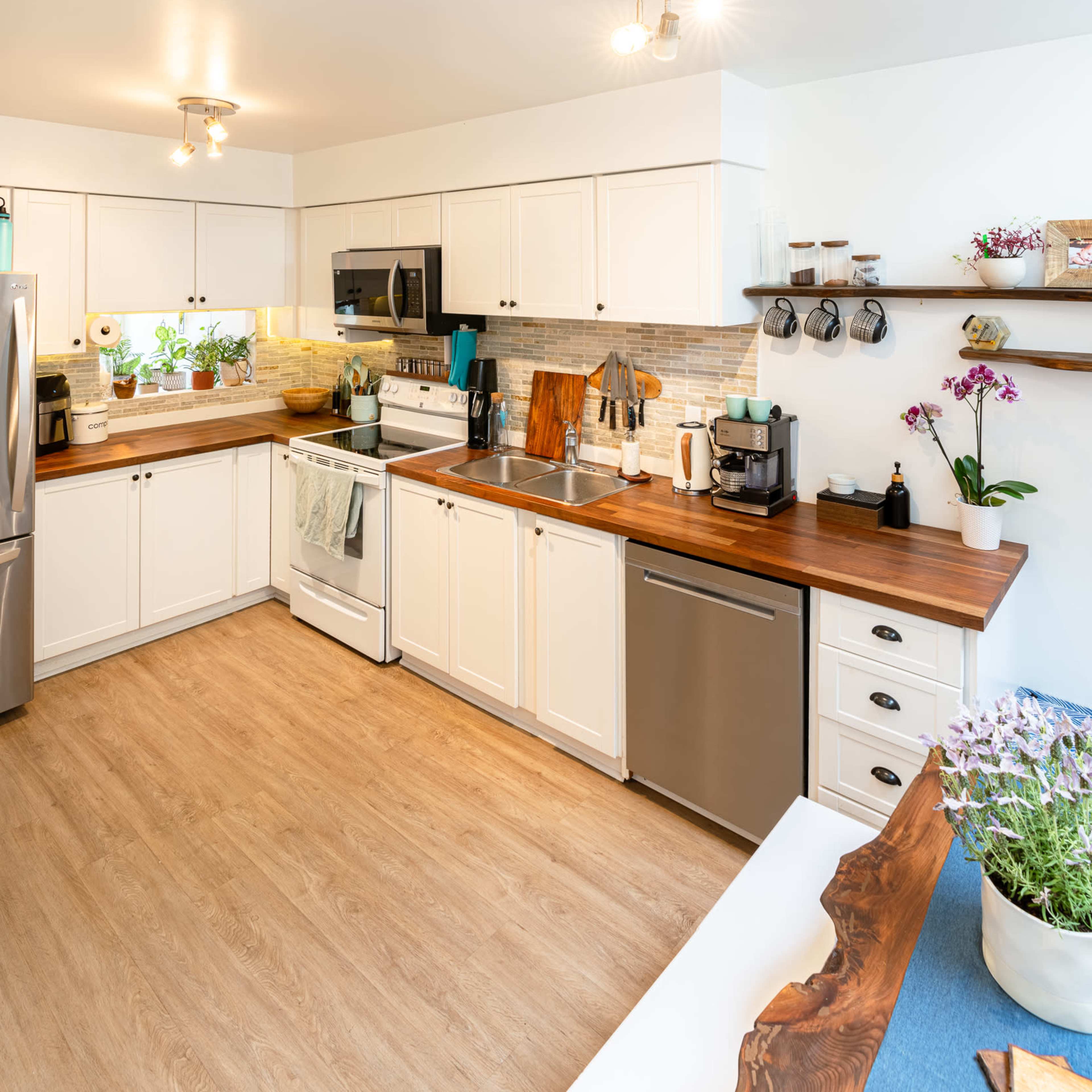 A modern kitchen features white cabinetry, stainless steel appliances, wooden countertops, and potted plants on the windowsill.