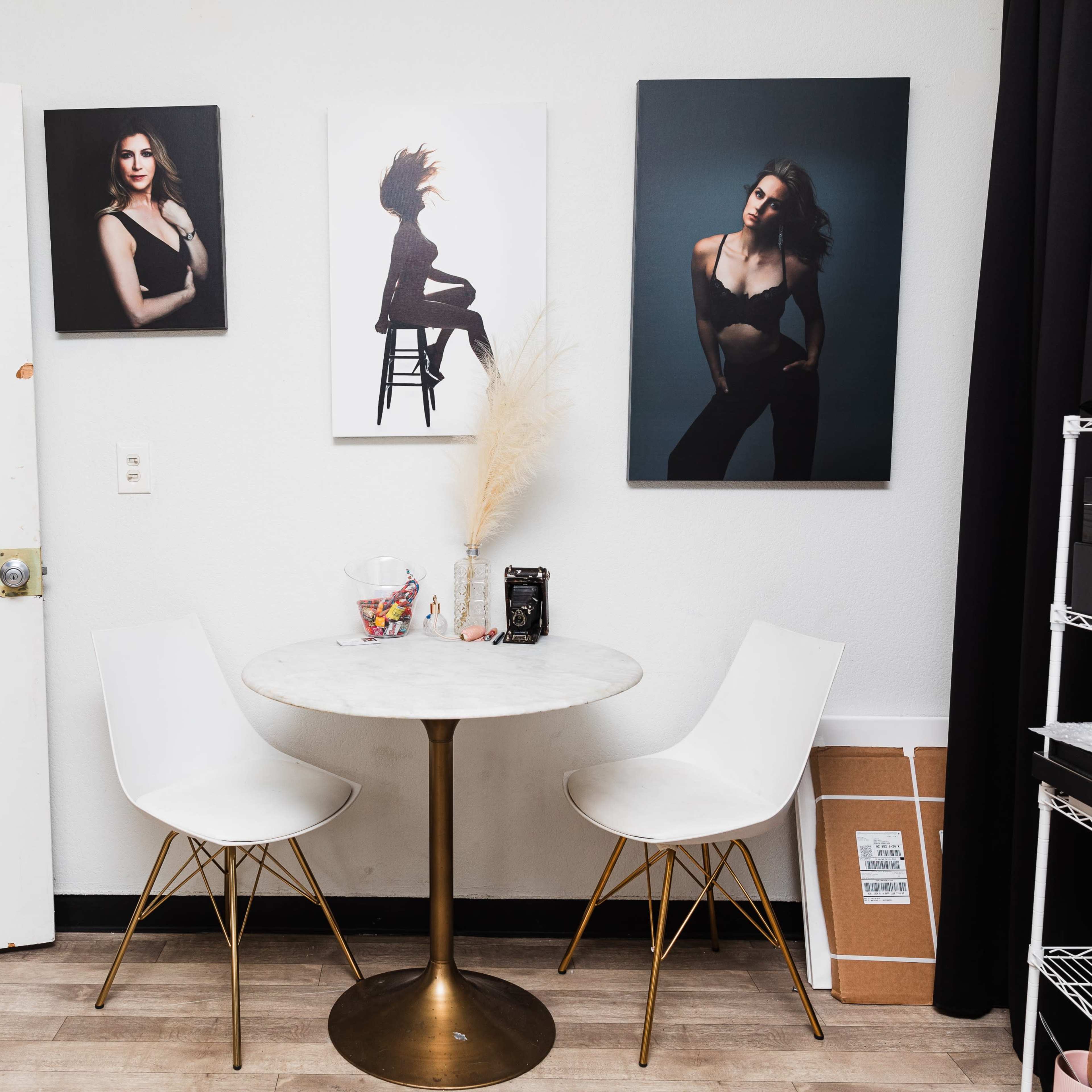 A small room features a round marble table with two white chairs, surrounded by three large framed photographs of a woman on the walls and a star emblem on the door.
