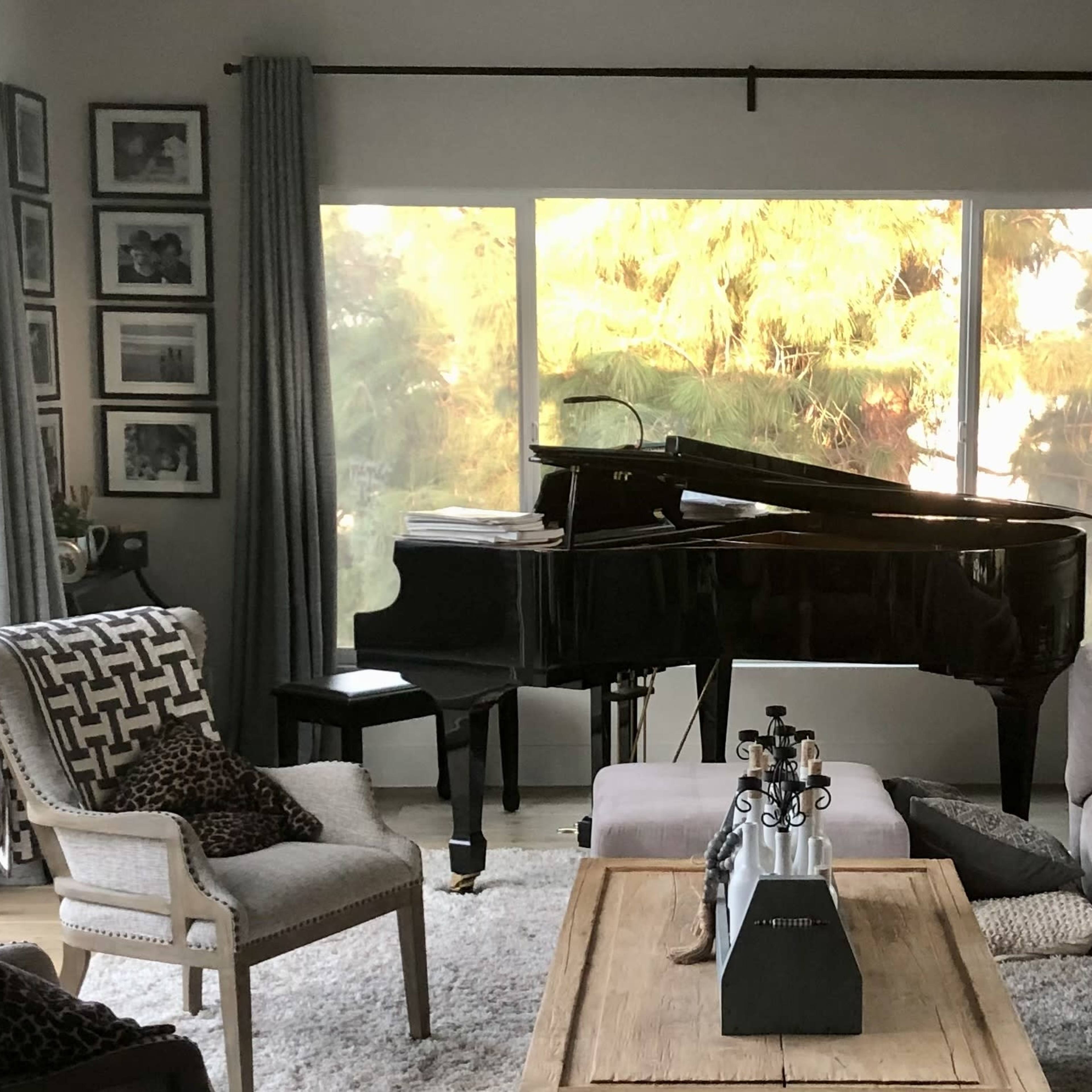 A grand piano sits in a well-lit living room beside a coffee table, with comfortable seating arranged around the space and large windows revealing a view of greenery outside.