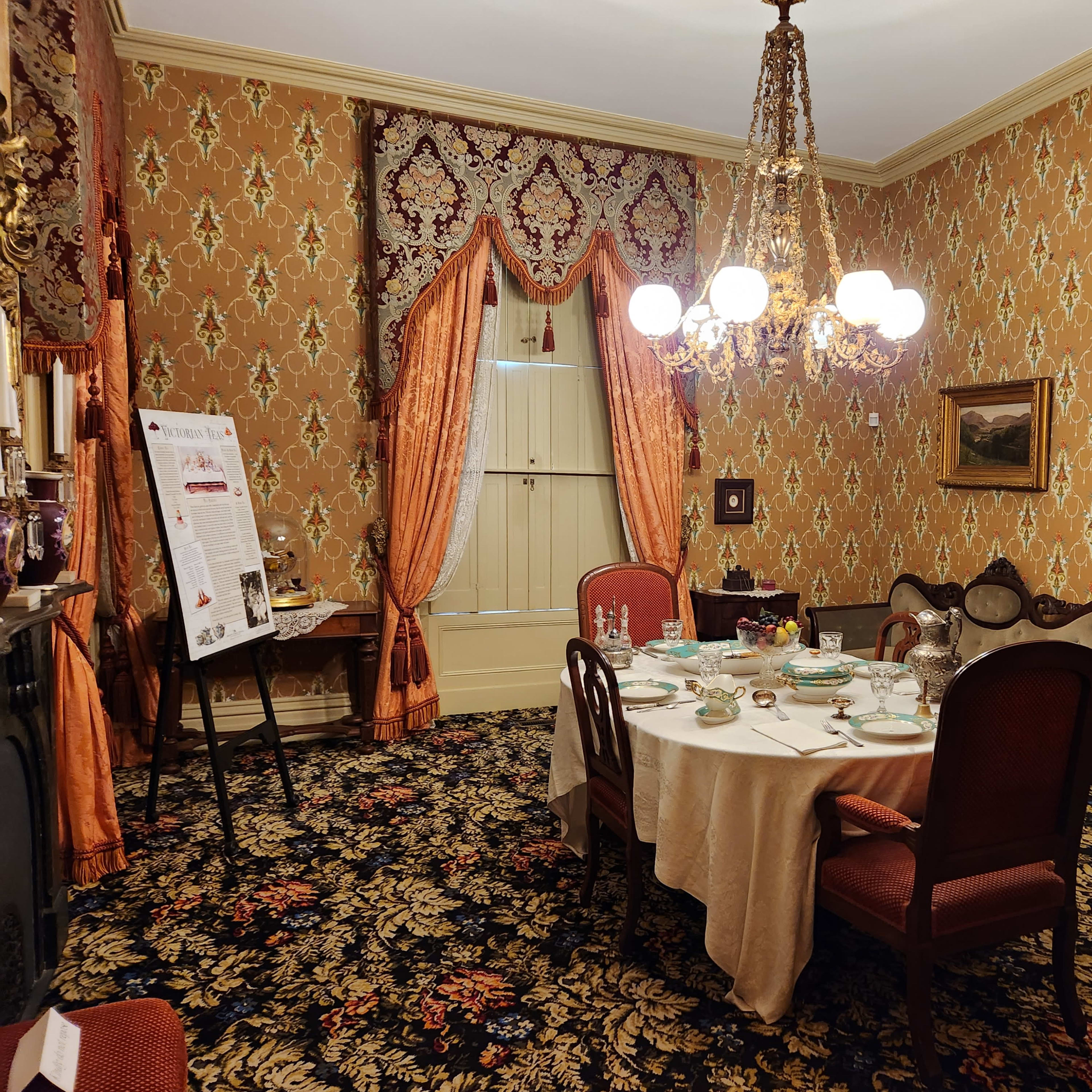 A formal dining room features a round table set with dishware, surrounded by wooden chairs, against a backdrop of ornate wallpaper and a chandelier.