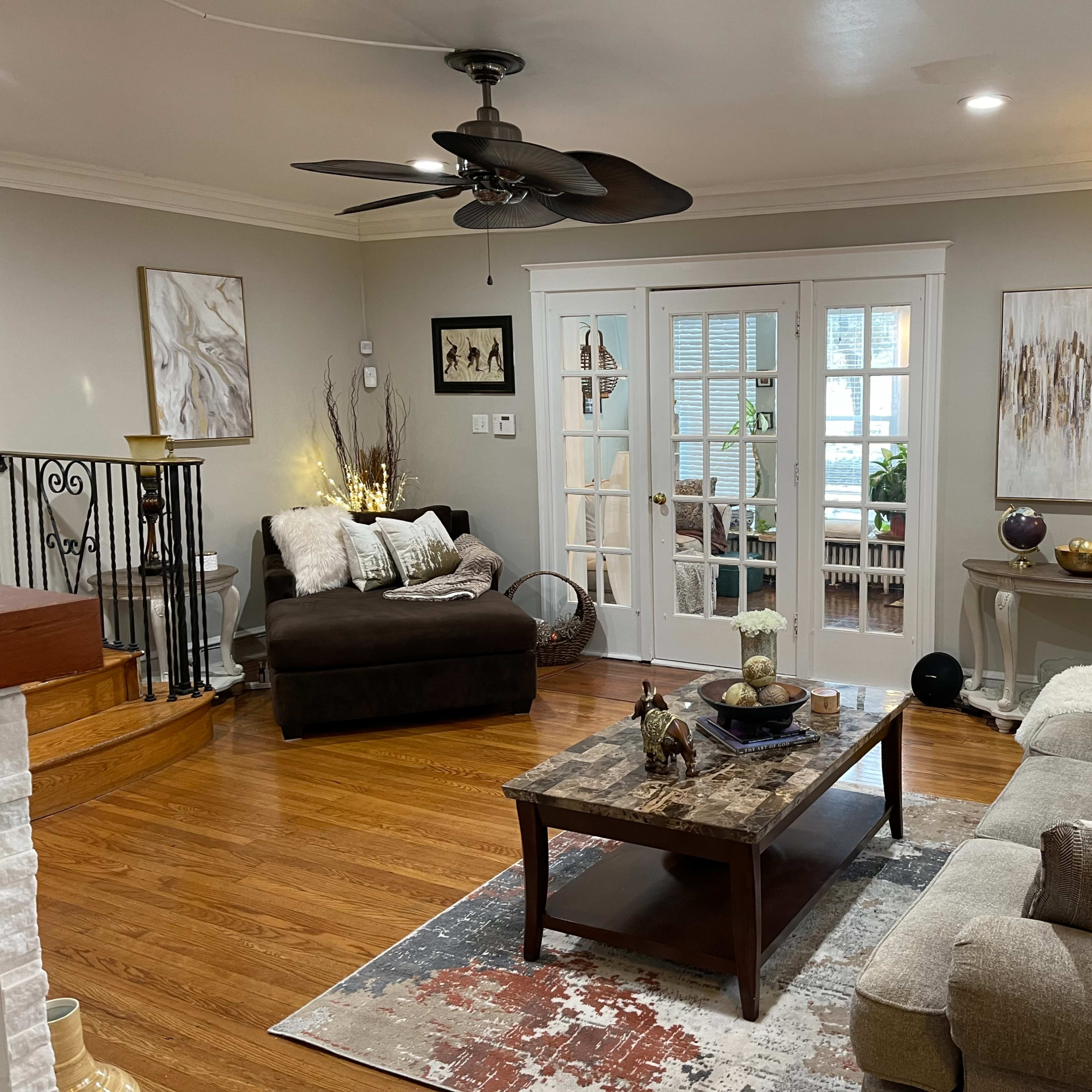 A cozy living room with a sofa, an armchair, a coffee table on a patterned rug, and large windows leading to another room.