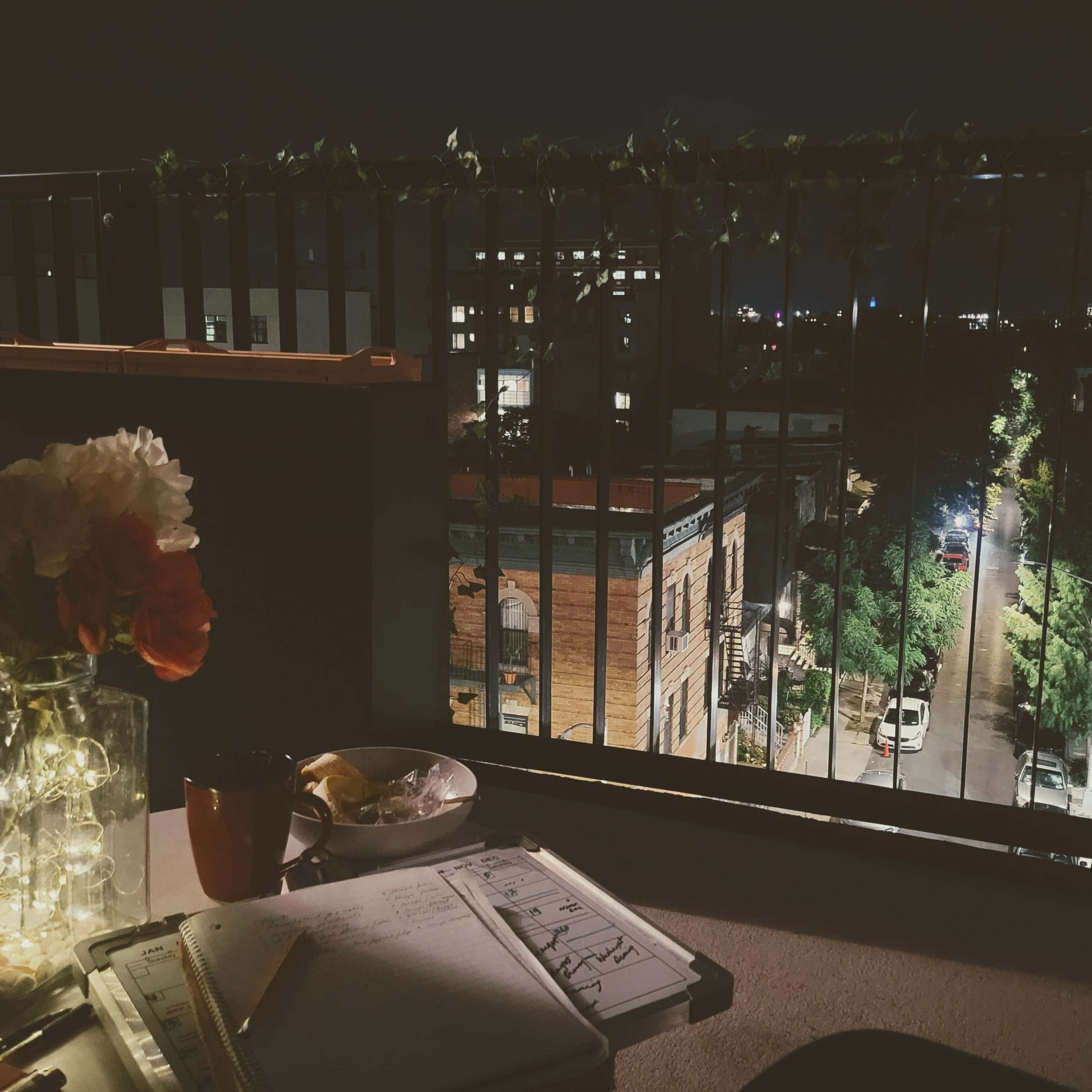 A balcony scene at night features a table with a notebook, a cup, a plate, and a vase of flowers, overlooking a lit street lined with trees and buildings.