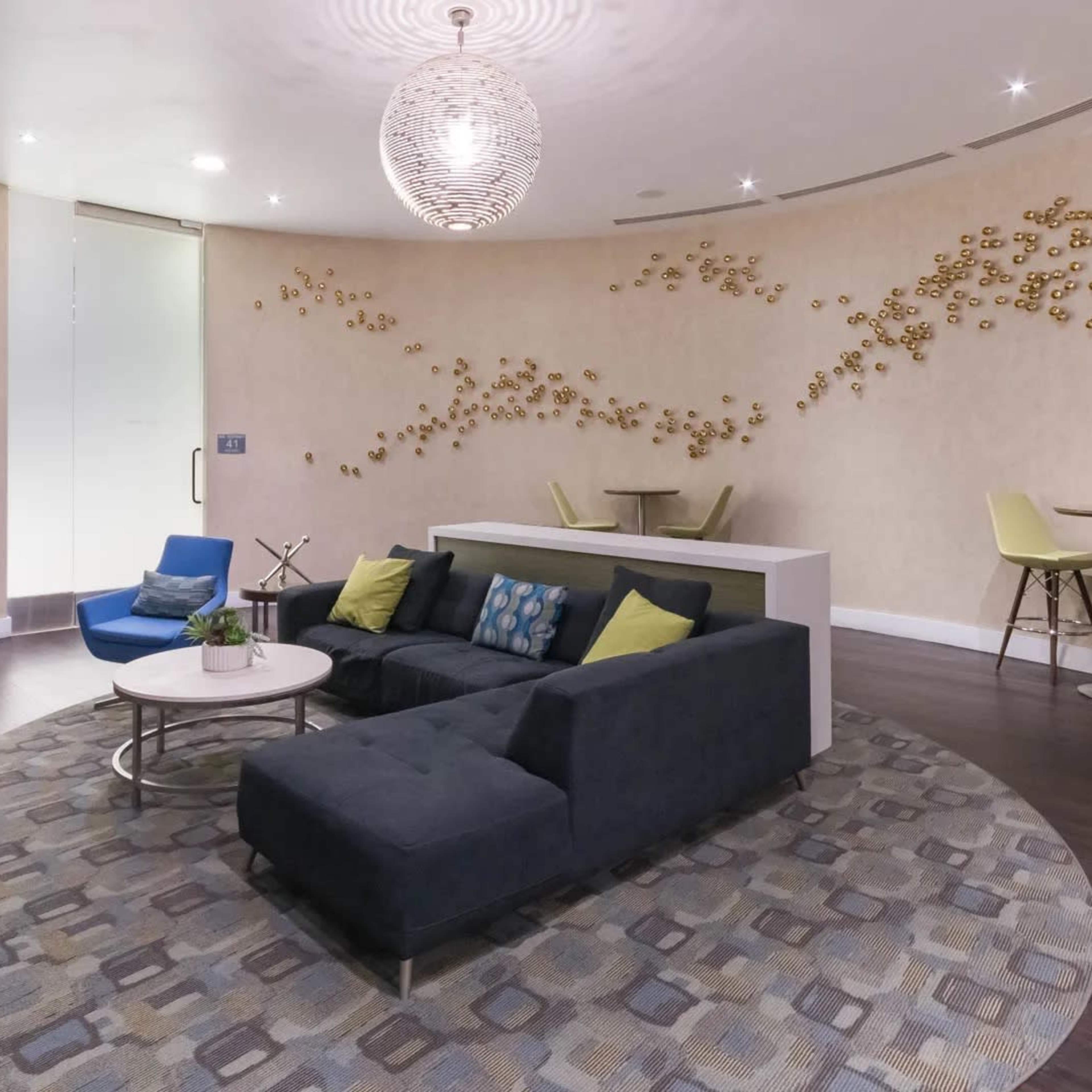 A modern lounge area with a circular layout featuring a dark sectional sofa, a round coffee table, decorative wall art, and light green high-top chairs against a textured wall.