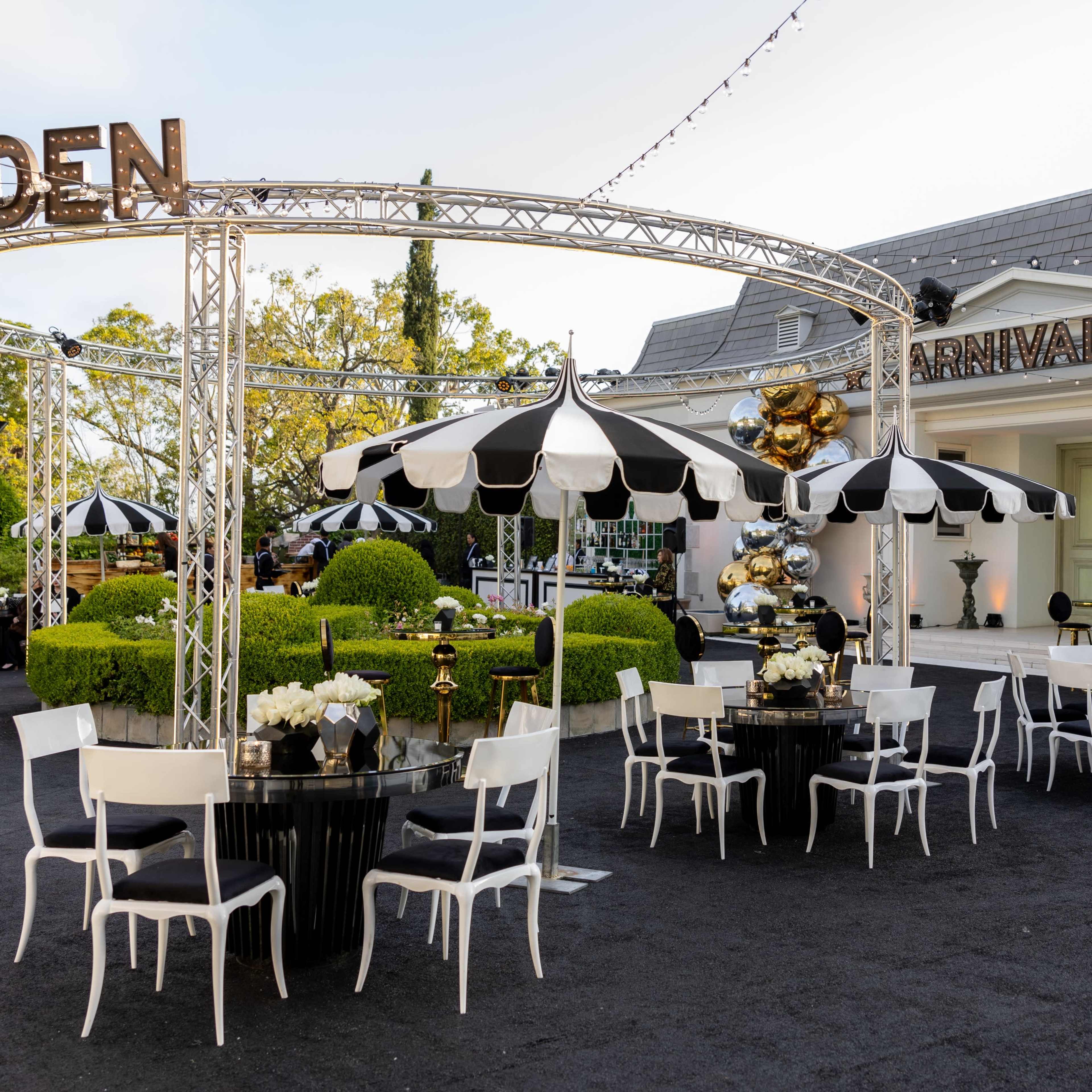 An outdoor event space featuring black and white striped umbrellas, circular tables adorned with white floral arrangements, and a backdrop of a building decorated with balloons.