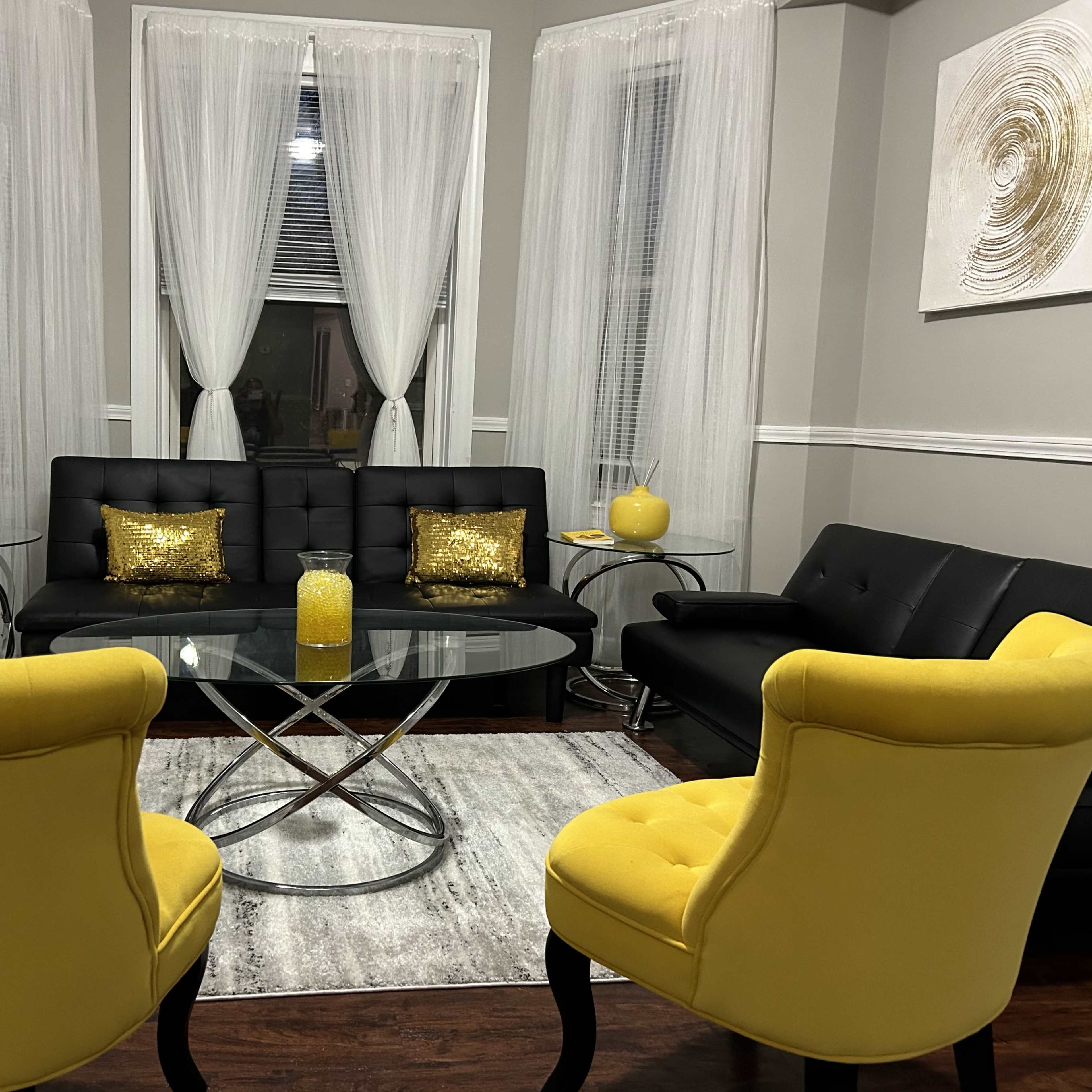 A cozy living room features a black leather couch, a glass coffee table, two yellow chairs, and sheer white curtains.