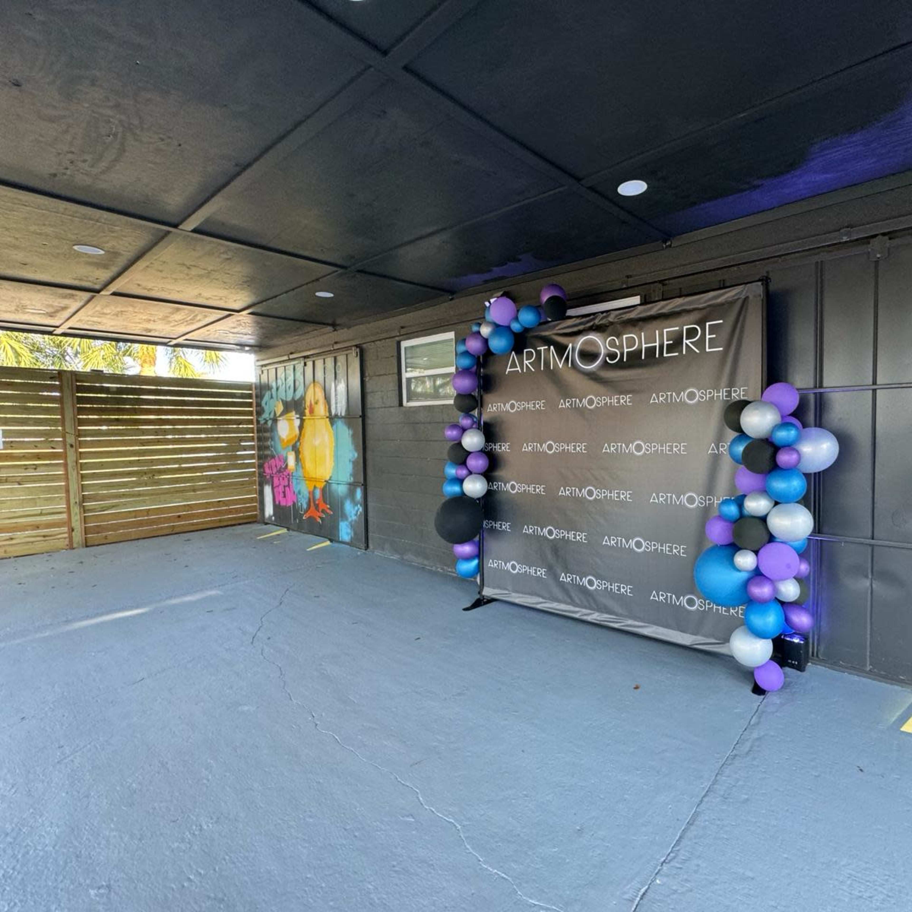 A decorated event space featuring a backdrop with the word "ARTMOSPHERE" and a cluster of purple, white, and blue balloons.