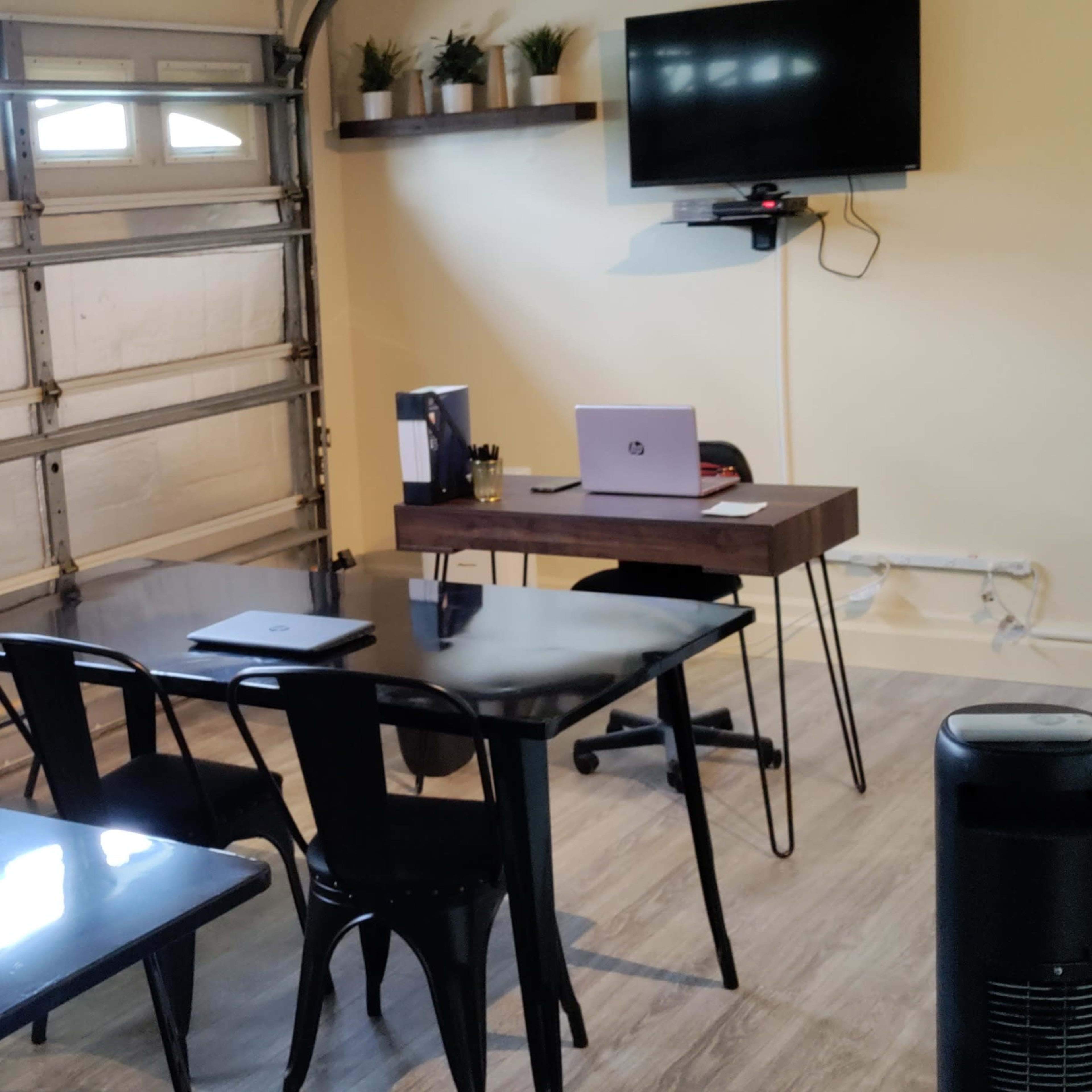 A garage transformed into a workspace, featuring a desk with a laptop, two tables, and a mounted television on the wall.