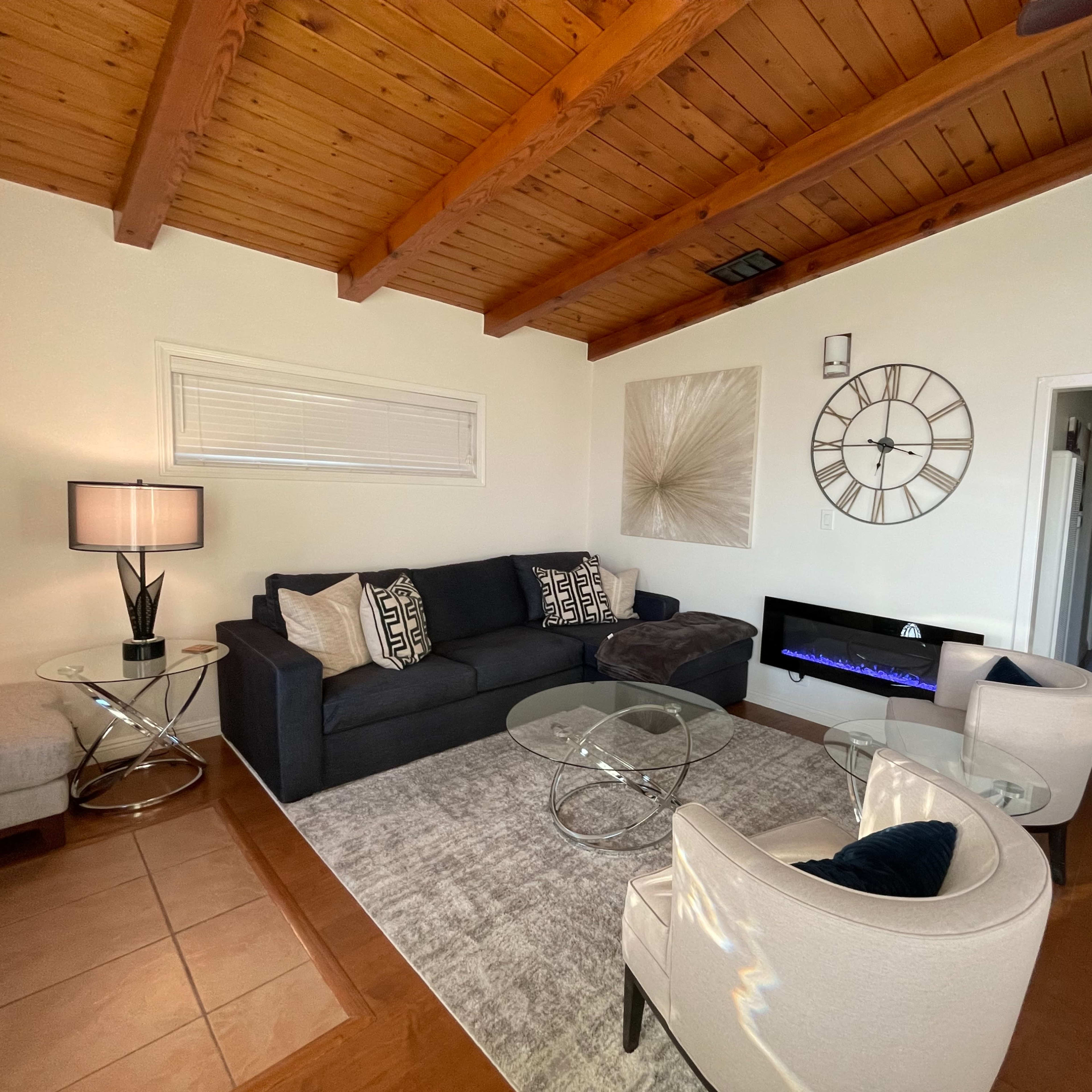 A living room with a dark sofa, a glass coffee table, two armchairs, and a modern electric fireplace beneath a wall clock.