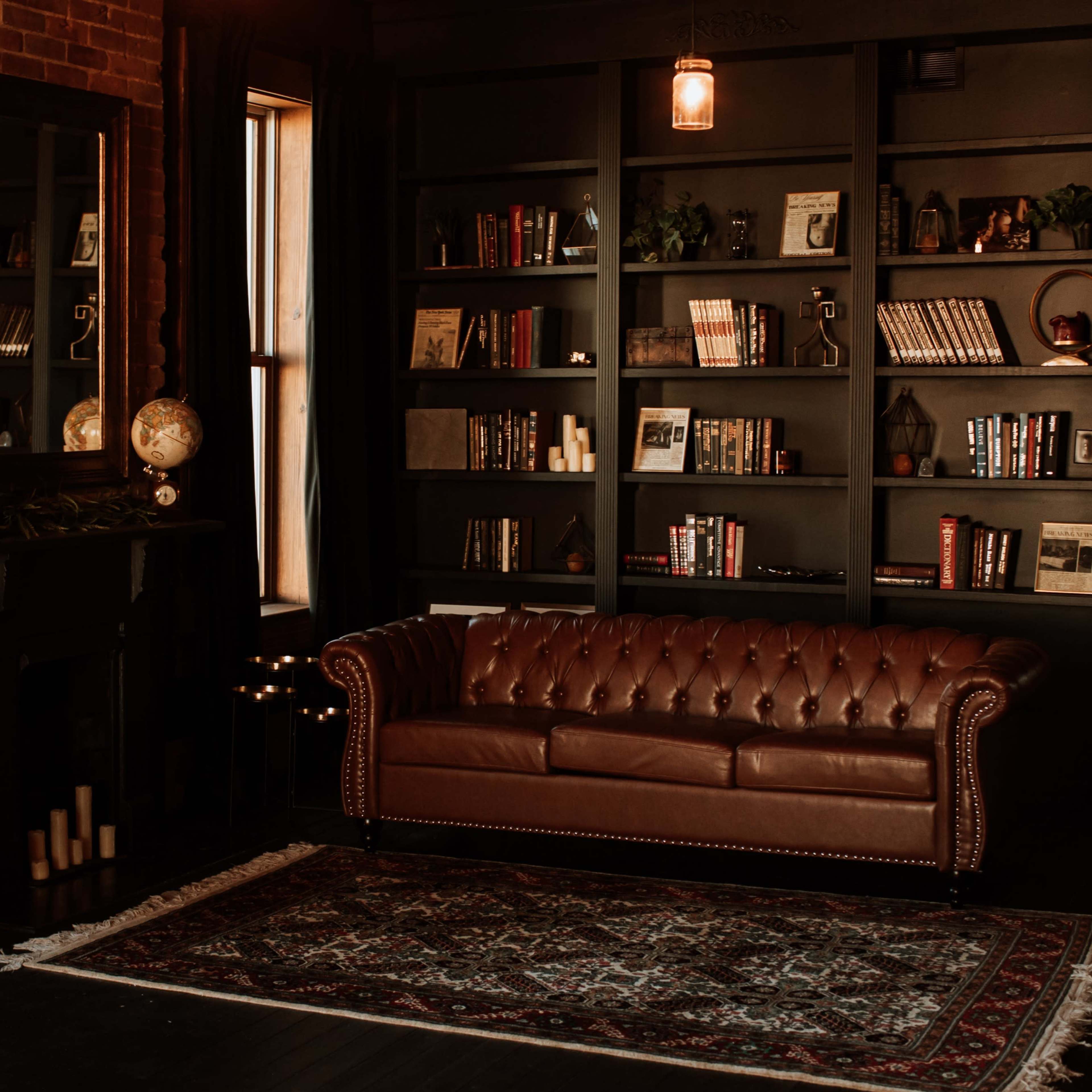 A cozy living room featuring a brown leather sofa, a dark bookshelf filled with books and decorative items, and a fireplace with candles.