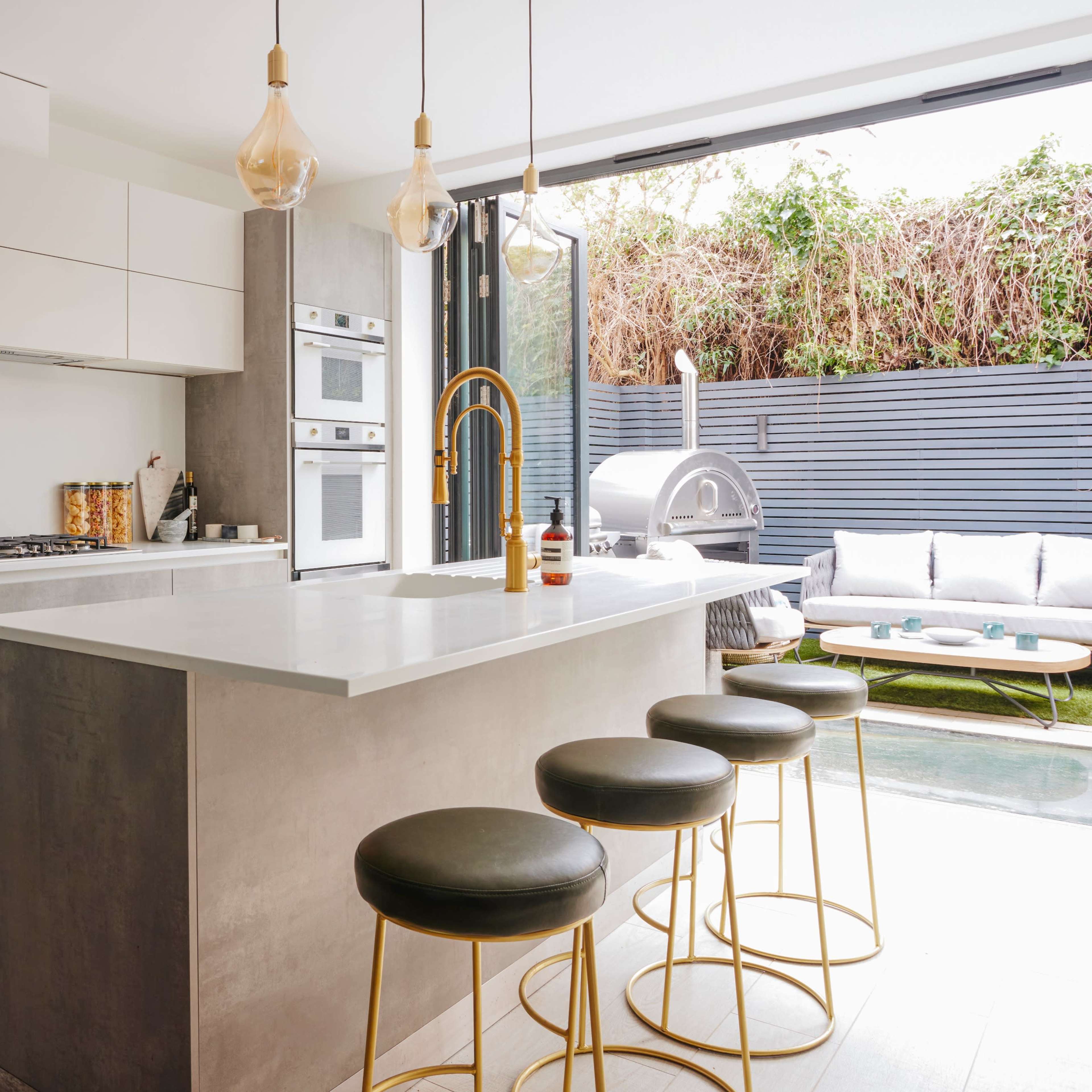 A modern kitchen with a central island, equipped with sleek cabinetry, stainless steel appliances, and four bar stools, leading to an outdoor seating area with a sofa and greenery.