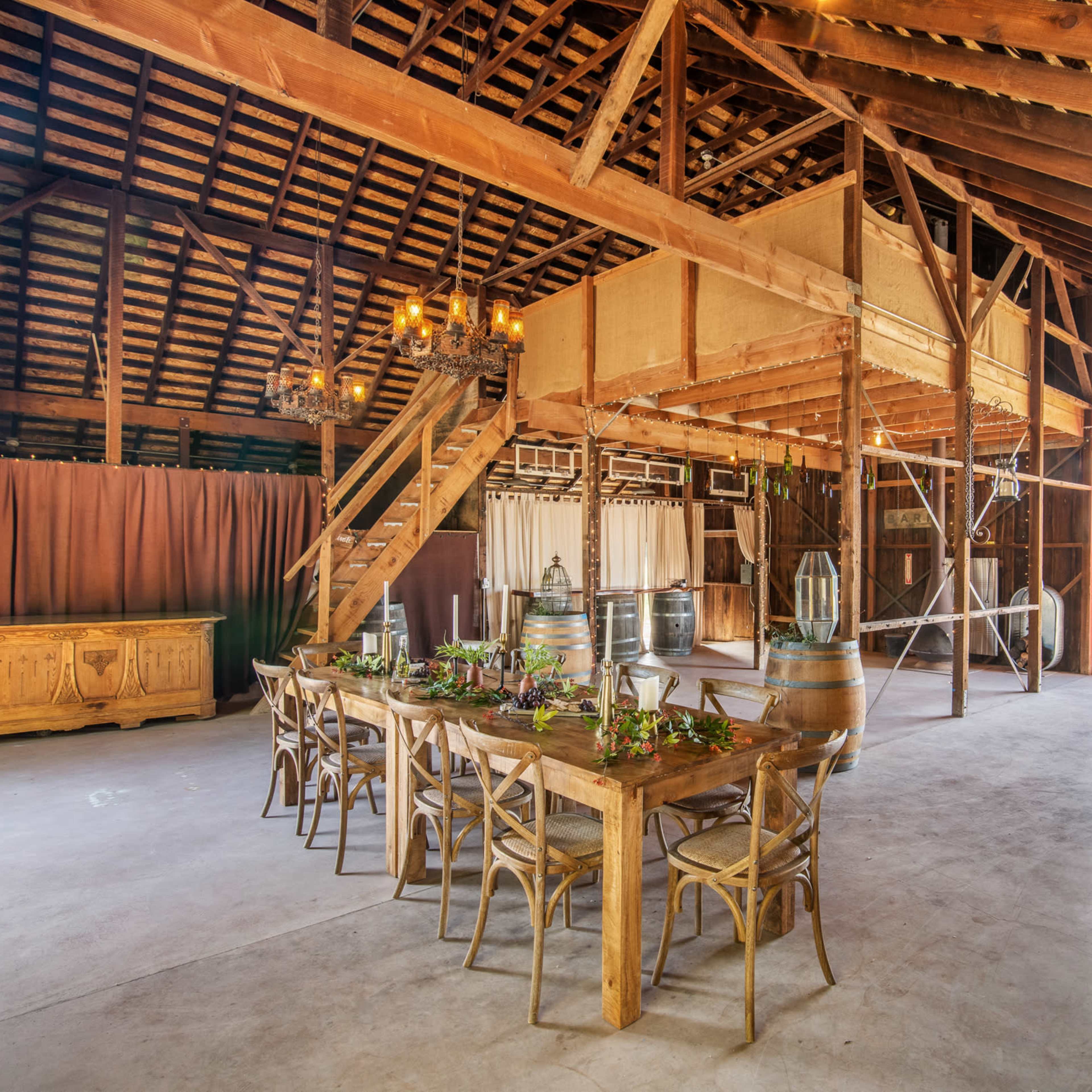The interior of a spacious barn features a wooden dining table set for a meal, with a staircase leading to a loft area and large windows allowing natural light to enter.