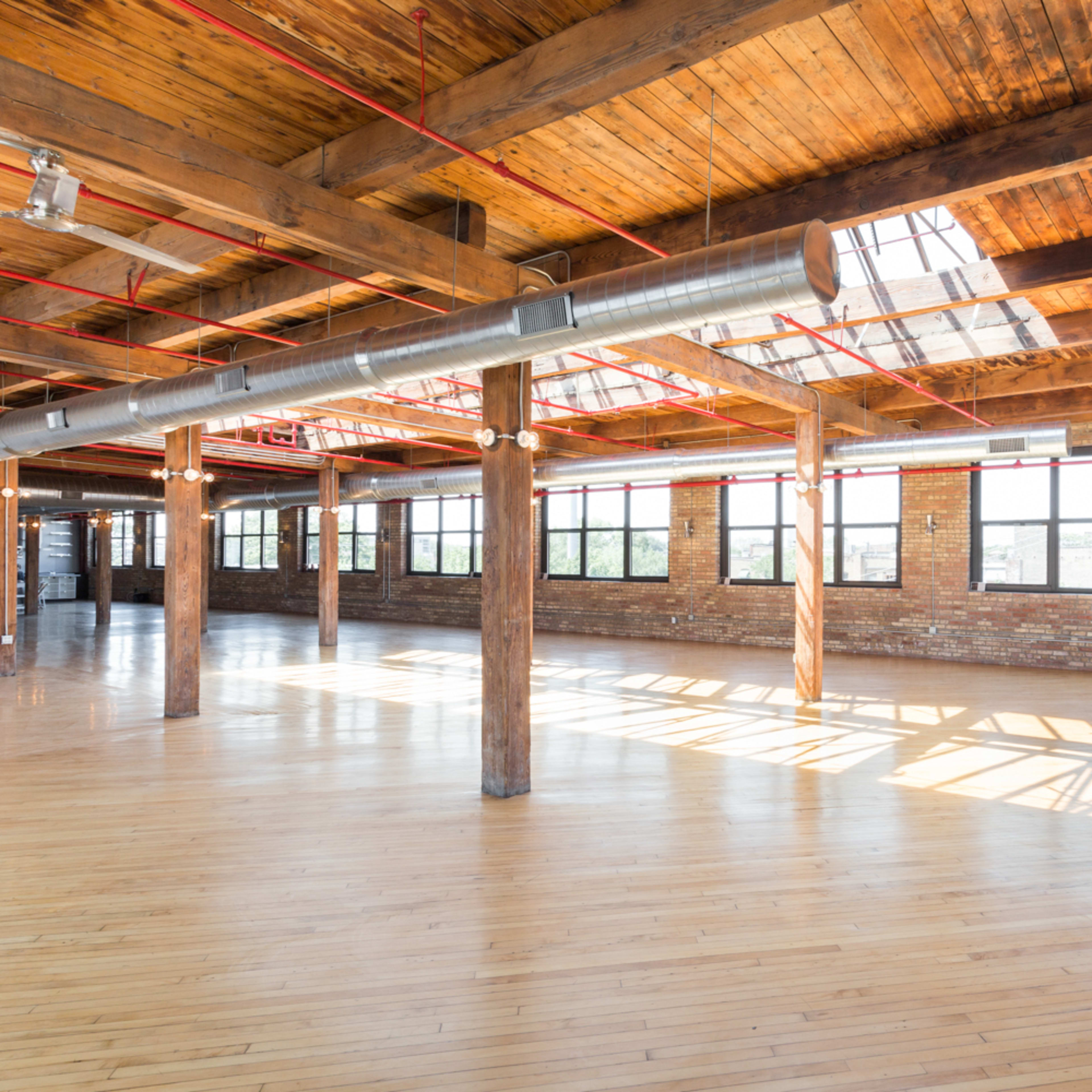 A spacious, sunlit industrial-style room with exposed wooden beams, brick walls, and large windows.