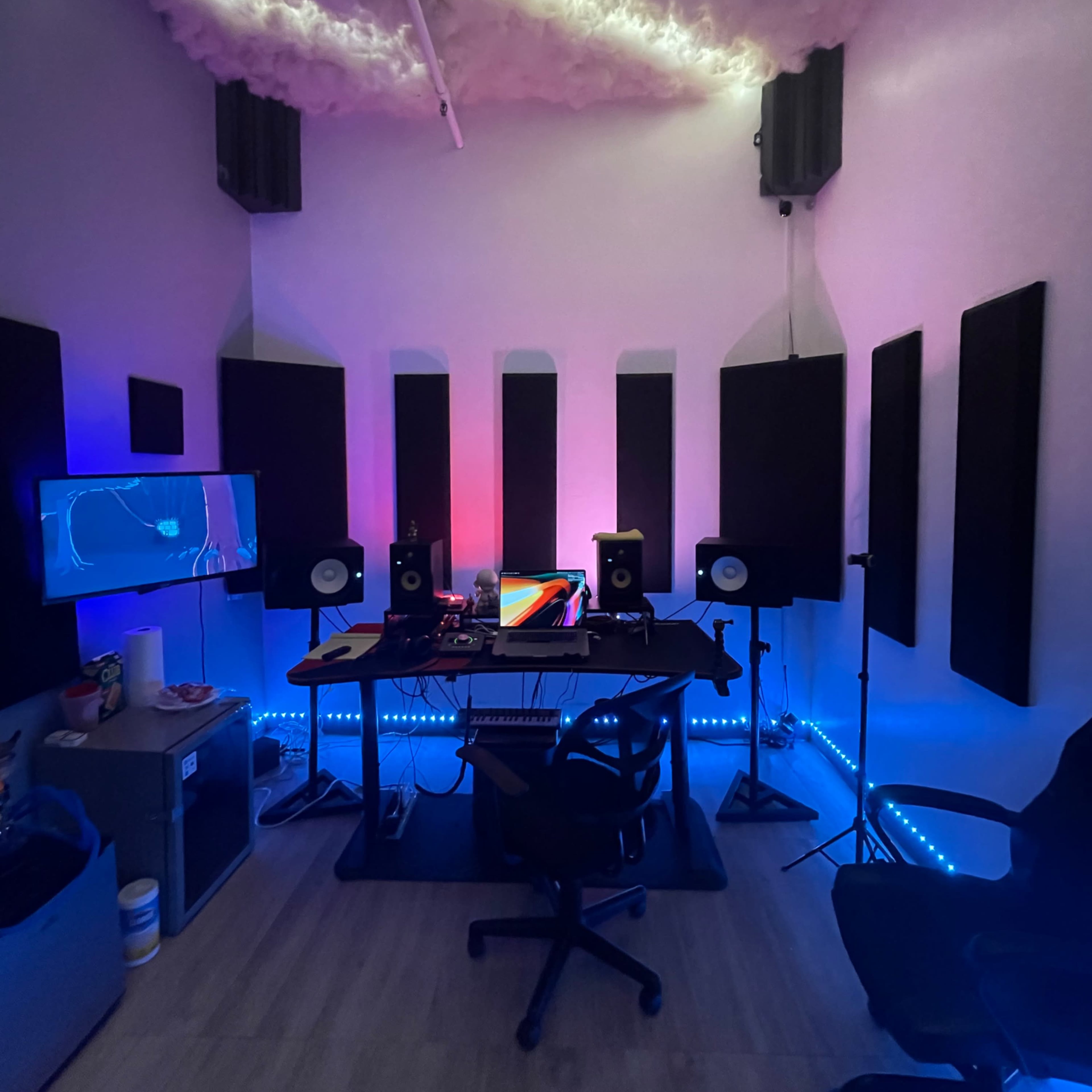 A music studio featuring a desk with audio equipment, monitors, a TV on the wall, and soundproof panels arranged around the room.