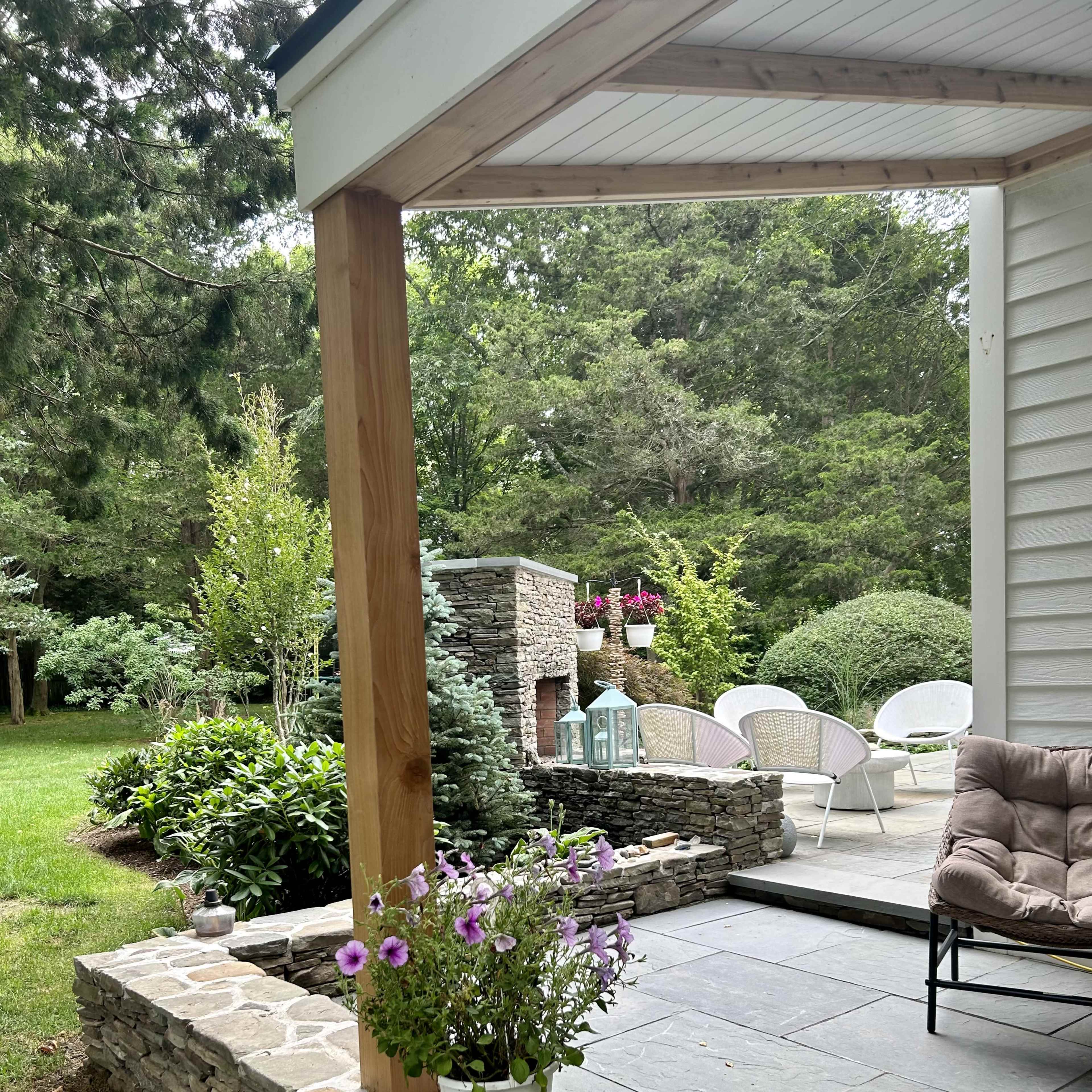 A covered patio with a stone fireplace overlooks a landscaped garden filled with greenery and outdoor seating.