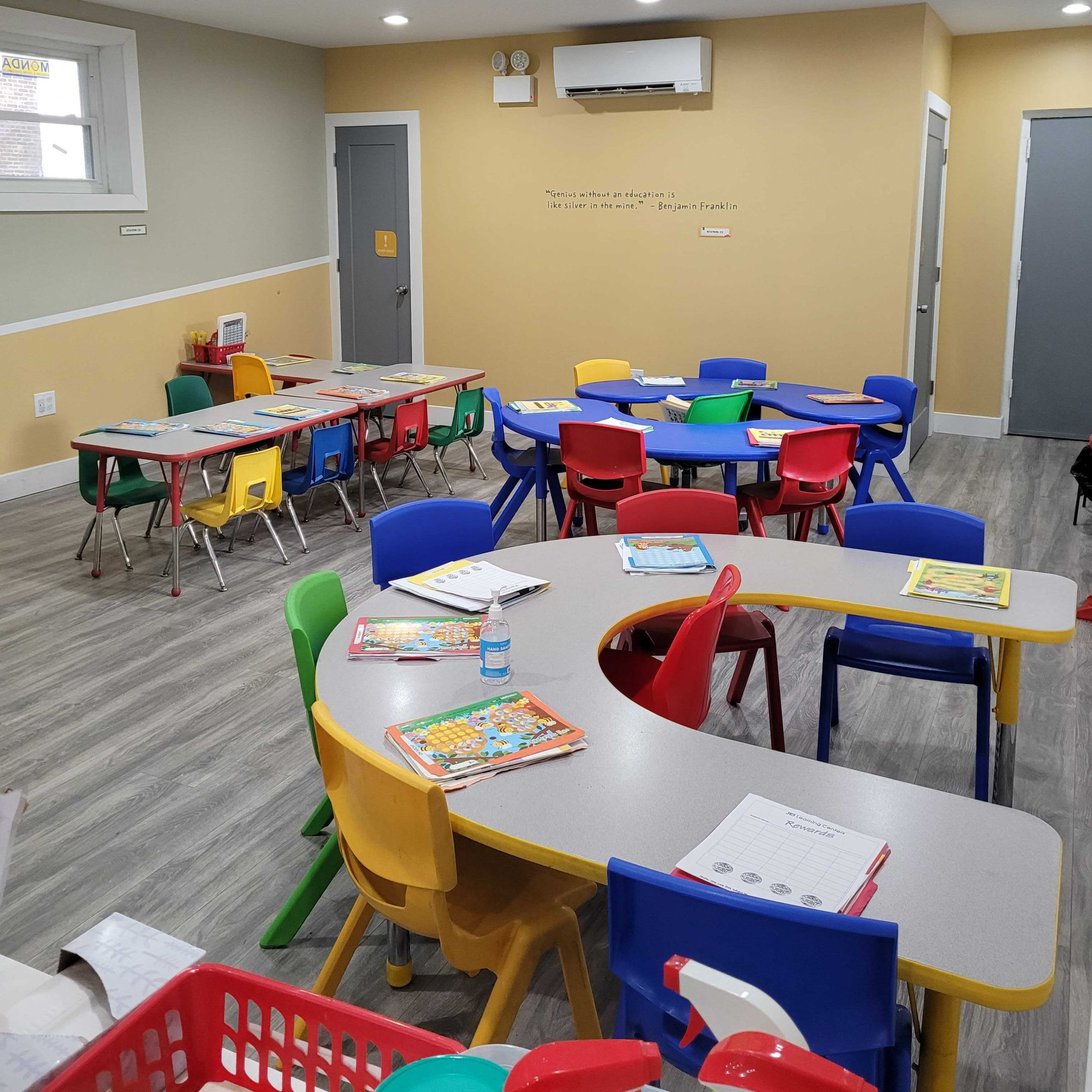 A brightly colored classroom with several round tables surrounded by chairs in various colors, along with educational materials scattered on the tables.