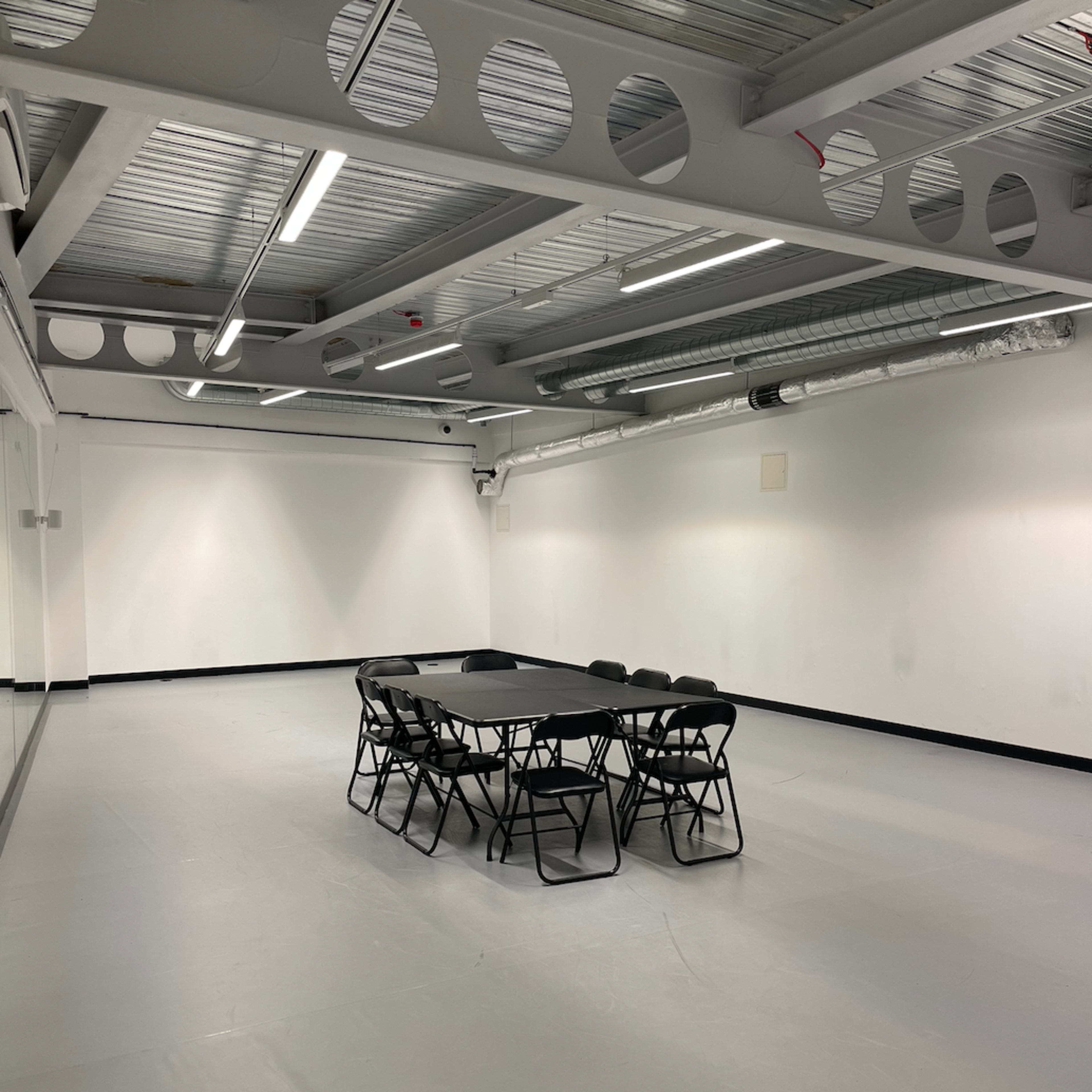 A large, empty room features a round table surrounded by black folding chairs on a smooth, gray floor.