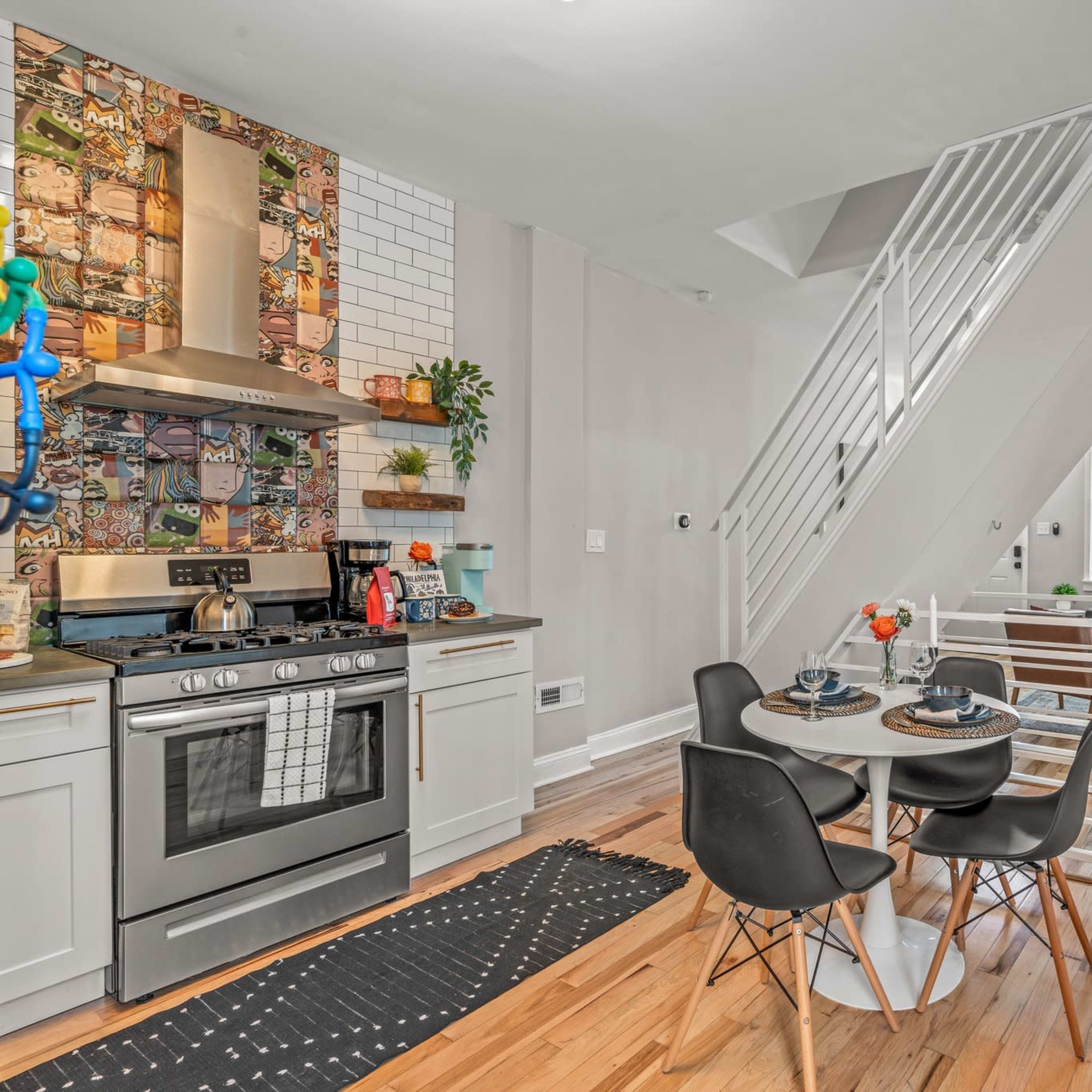 A modern kitchen area features a round dining table with black chairs, a stainless steel stove, and a tiled backsplash, leading to a living room with large windows and a sectional couch.
