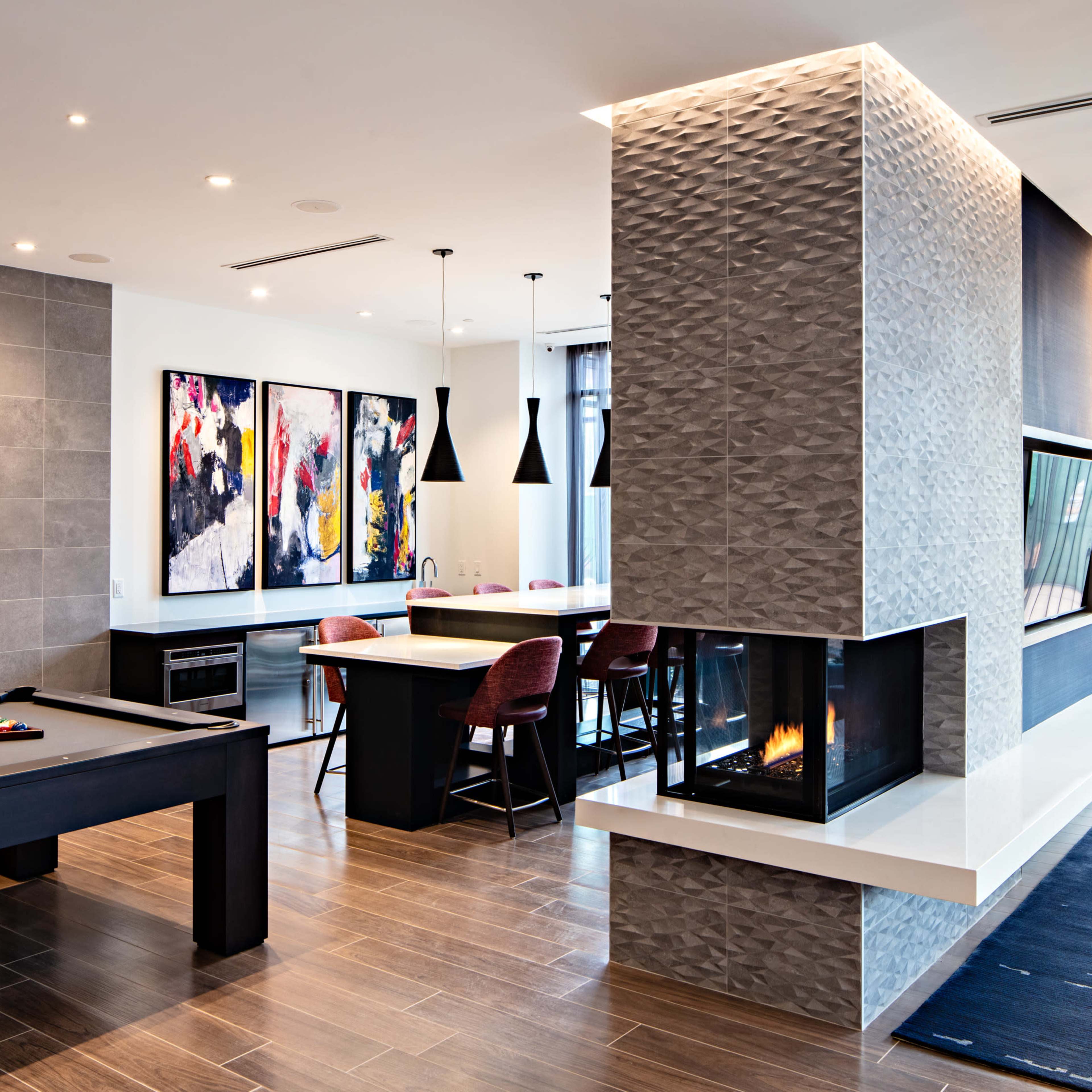 A modern living space featuring a pool table, a dining area with bar stools, a two-sided fireplace, and large windows with natural light.