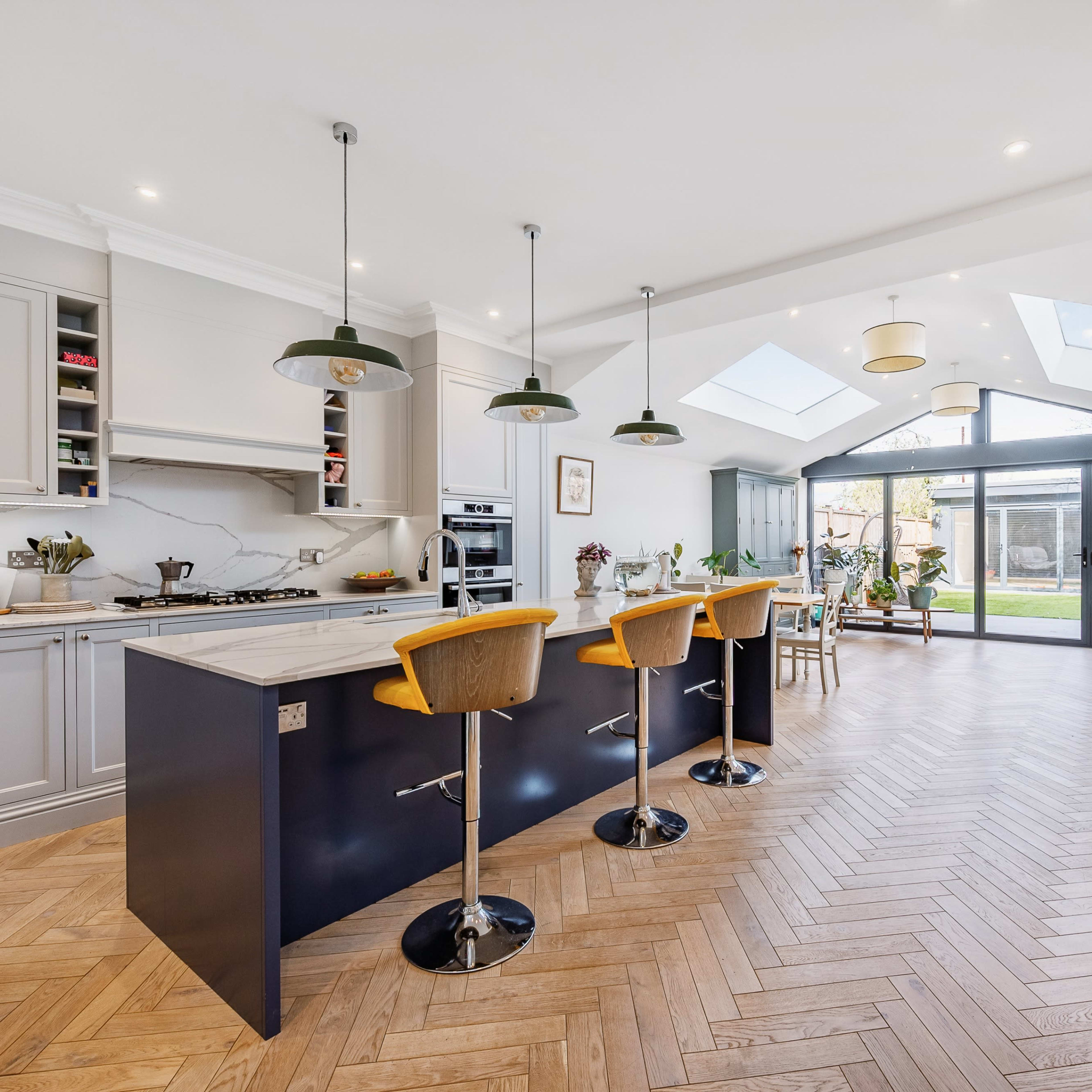 A modern kitchen features a central island with bar stools, sleek cabinetry, and large windows overlooking a backyard patio.