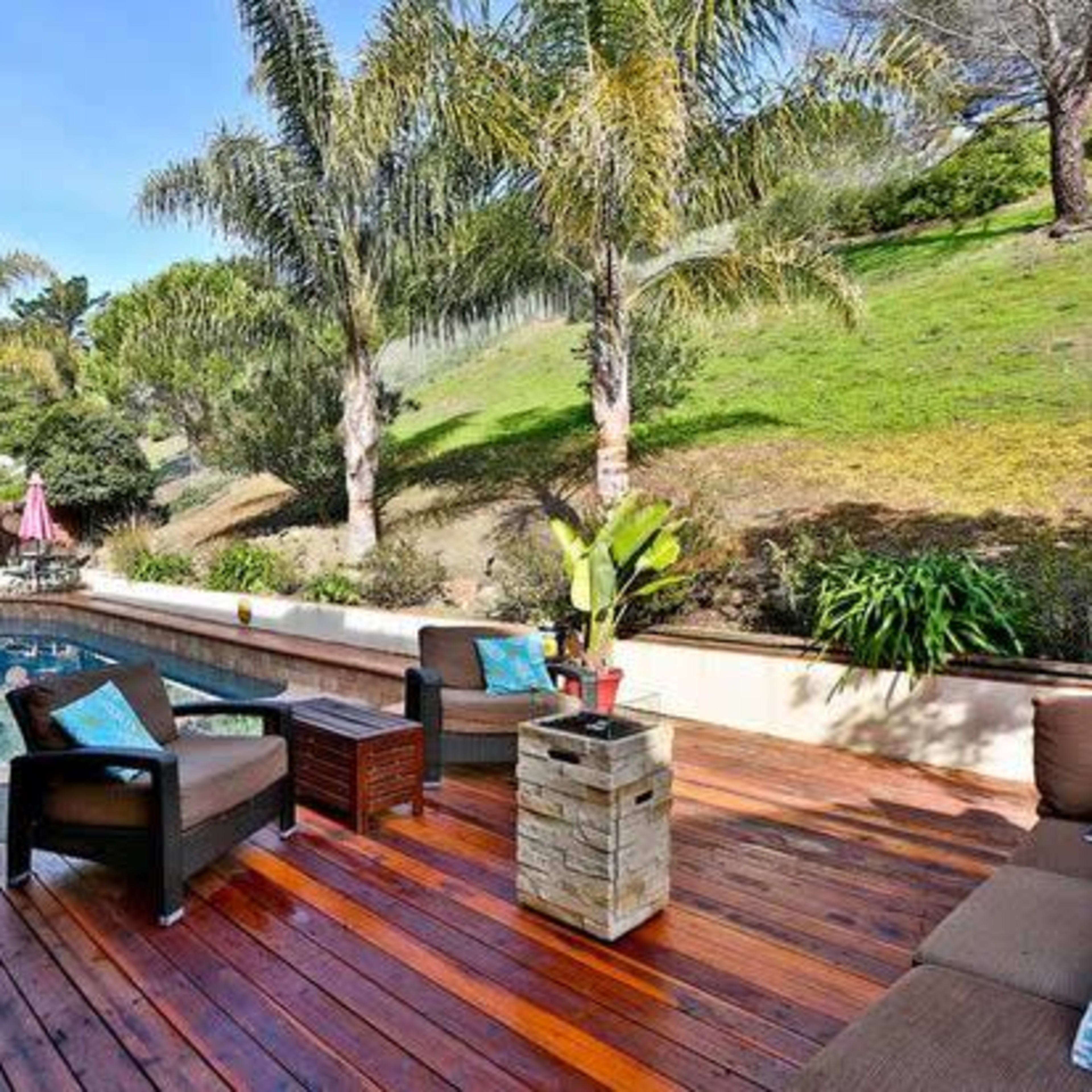 A backyard patio features a swimming pool surrounded by palm trees and wooden furniture.