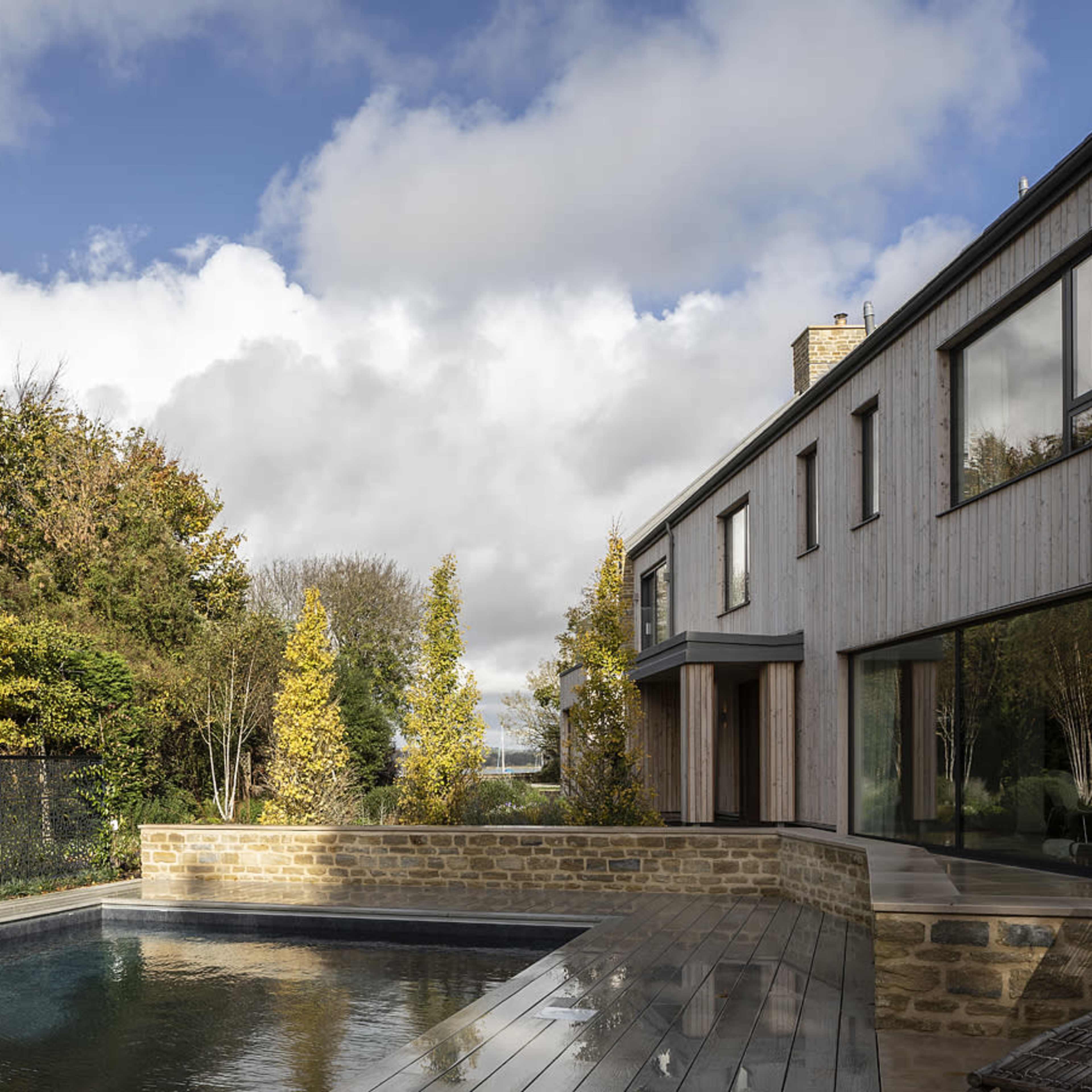 A modern two-story house with large windows, situated beside a rectangular swimming pool and surrounded by greenery under a cloudy sky.