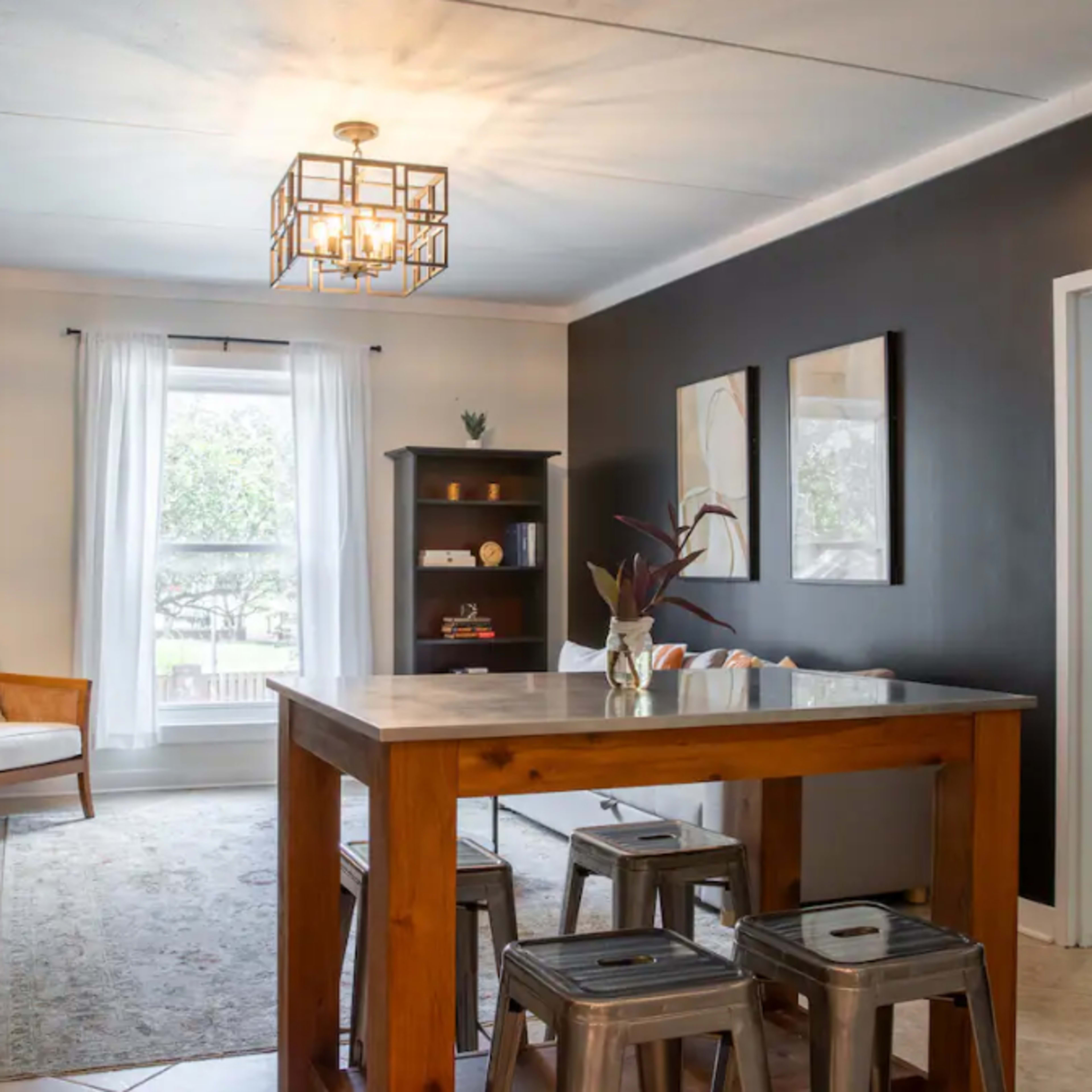 A living area with a wooden dining table, metal stools, a television, and a bookshelf, set against dark walls and featuring a large window with sheer curtains.