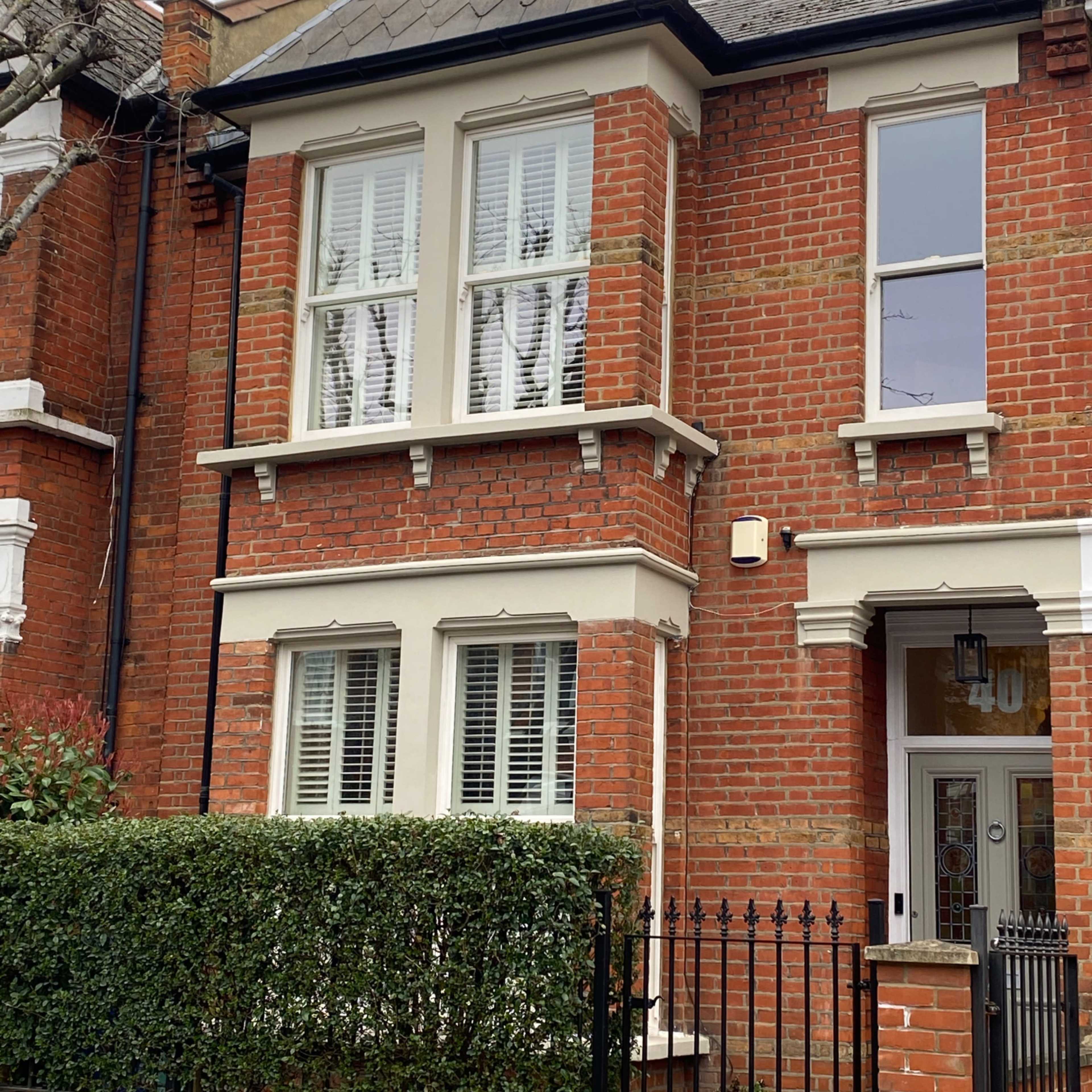 A brick townhouse with white window shutters and a small garden enclosed by a black wrought-iron fence.