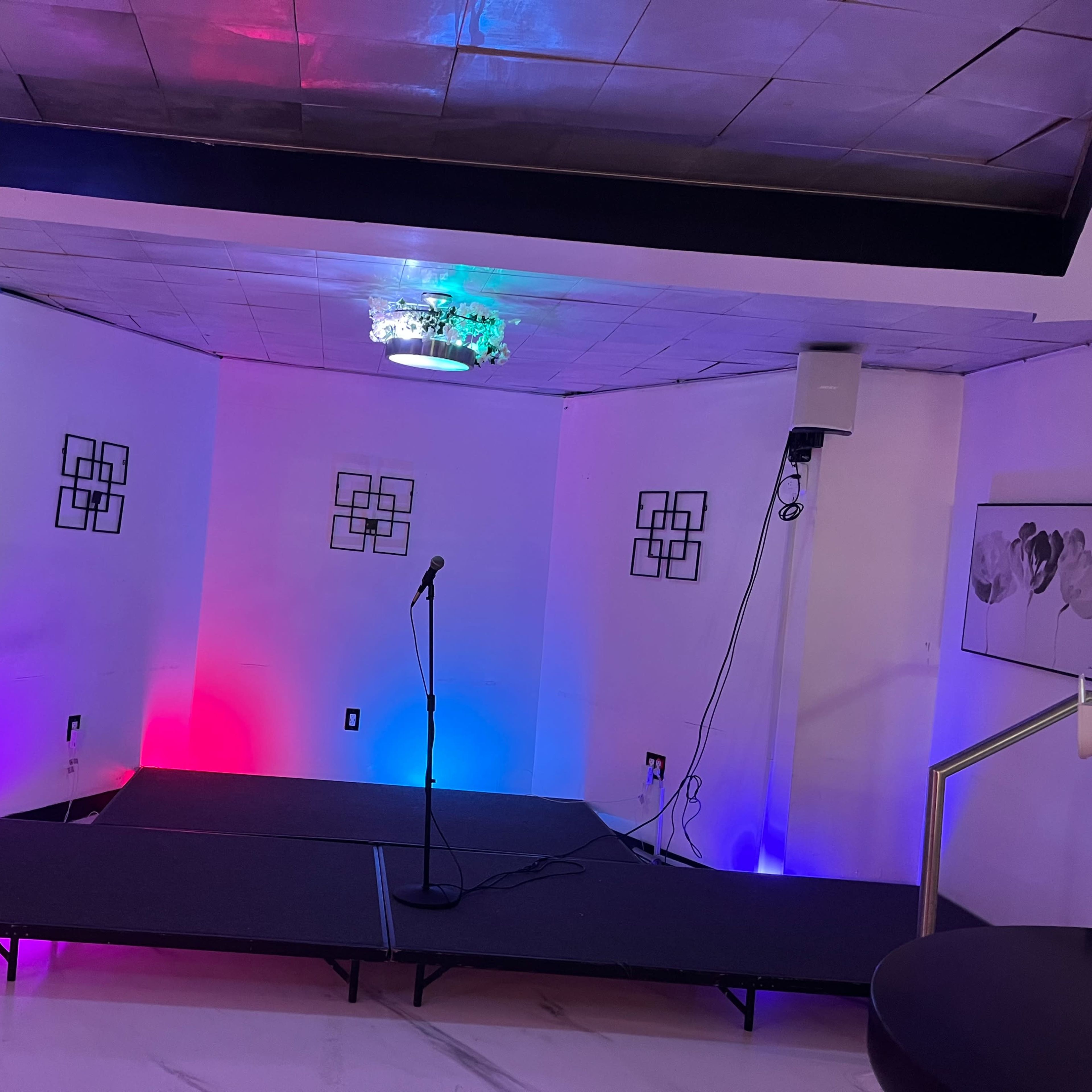 A small stage with a microphone in front, illuminated by colored lights, set against a white wall adorned with geometric decorations.