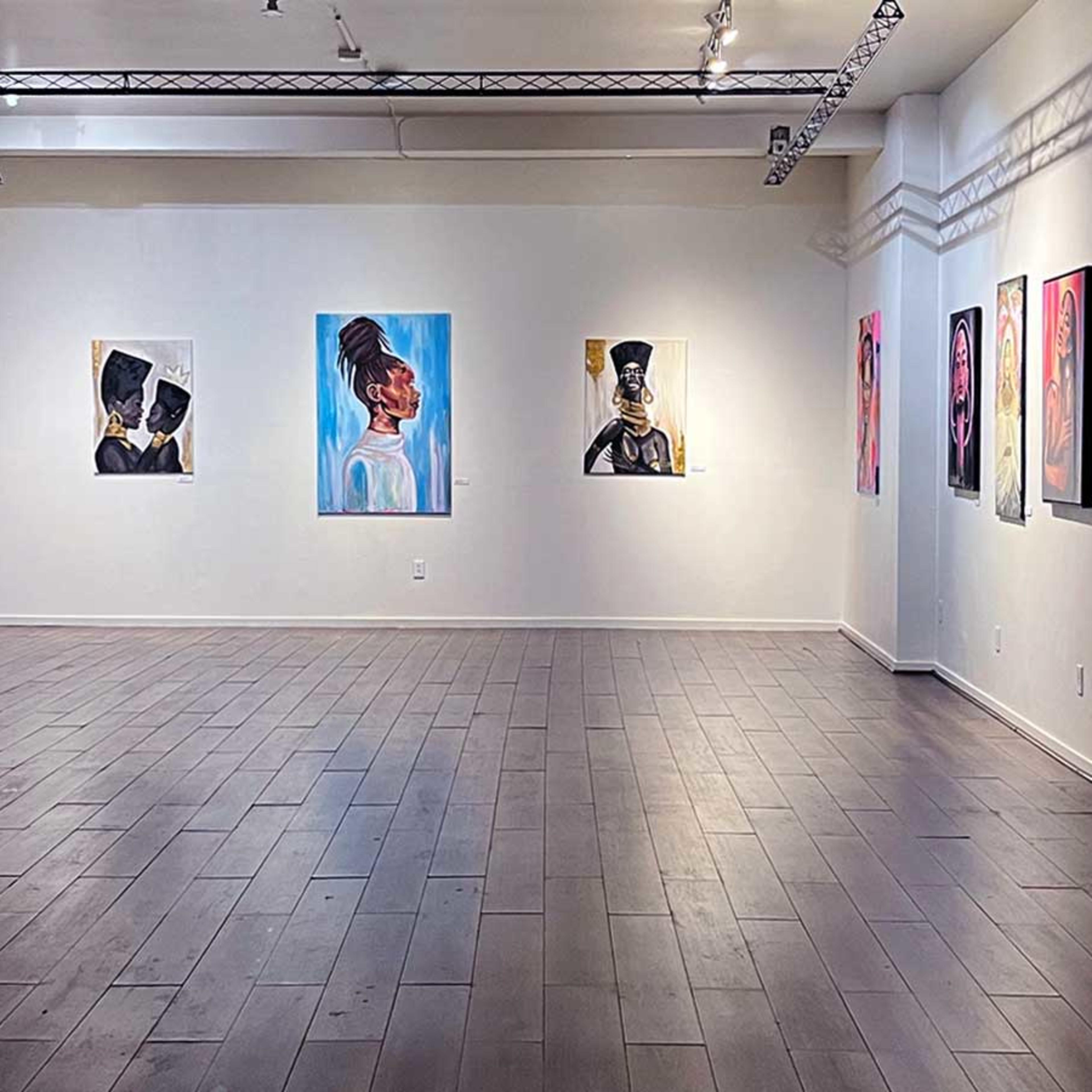 A spacious art gallery featuring various paintings displayed on the white walls, with exposed brick on one side and polished wooden flooring.