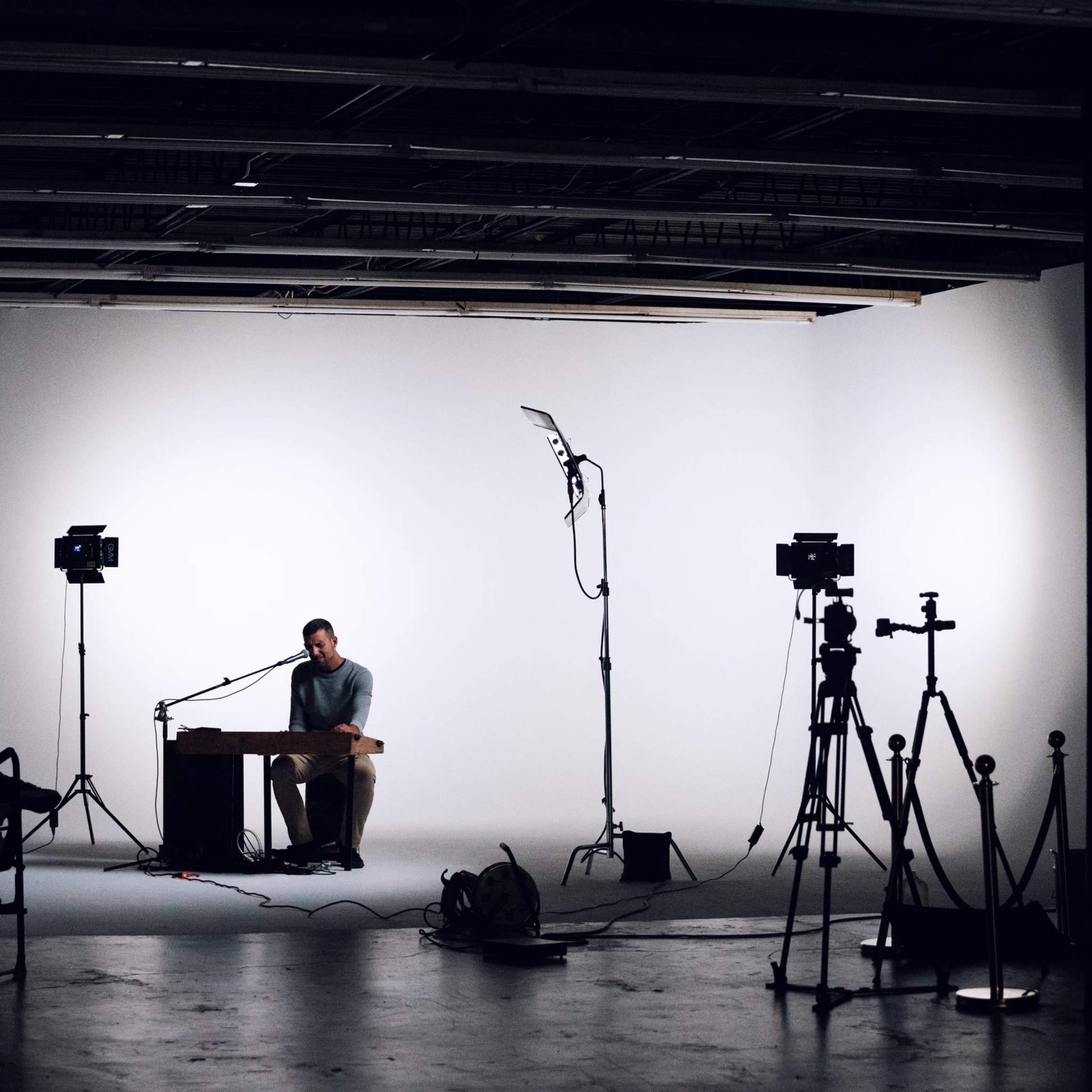 A musician sits at a table in a dimly lit studio with cameras and lighting equipment set up around him.